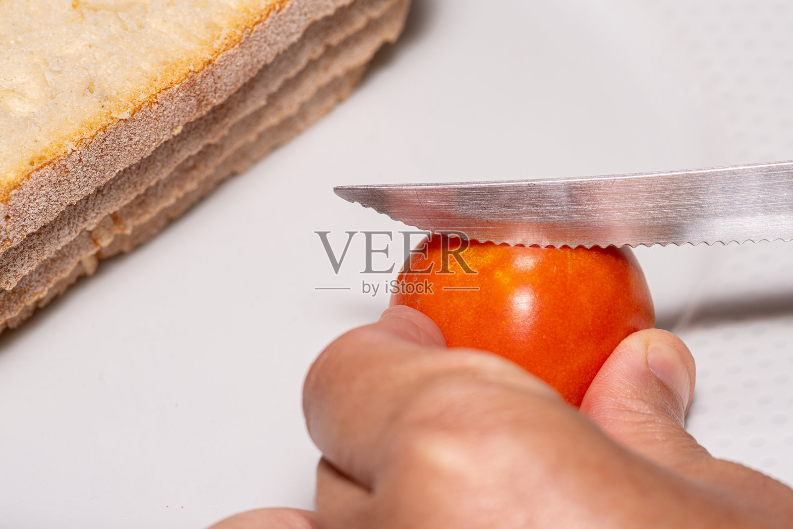 一只手握着番茄，刀子切割番茄，然后将其涂抹在一片面包上。照片摄影图片