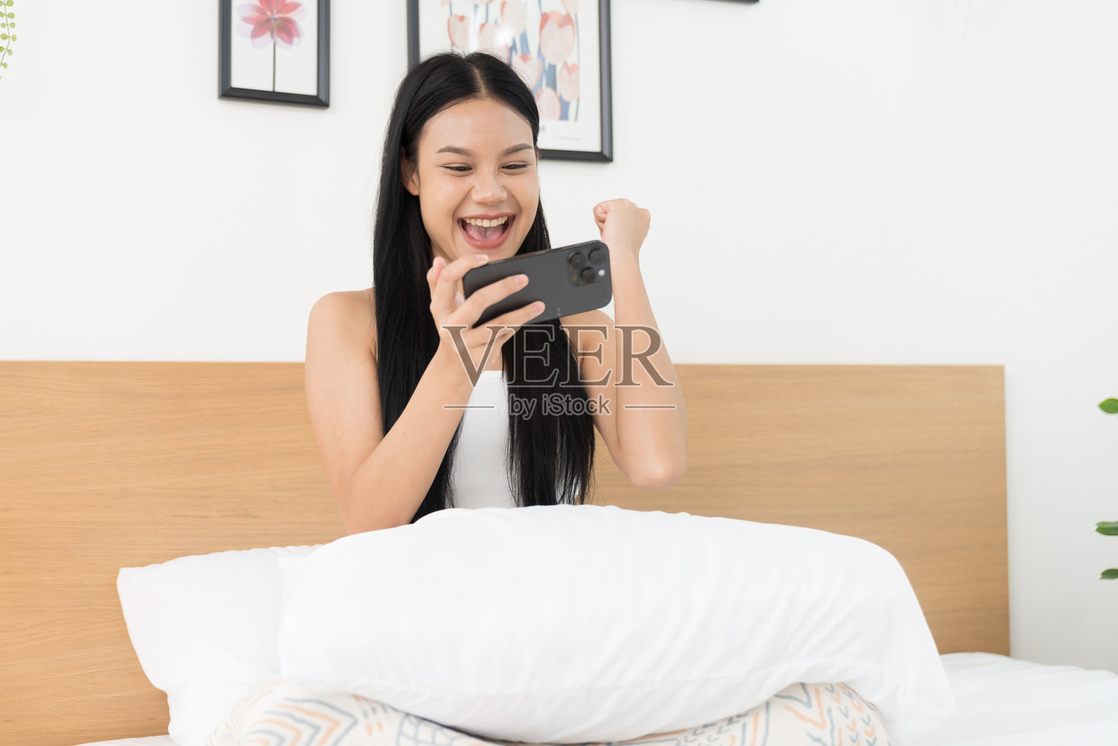 愉快的年轻亚洲女性在卧室的床上玩手机游戏照片摄影图片