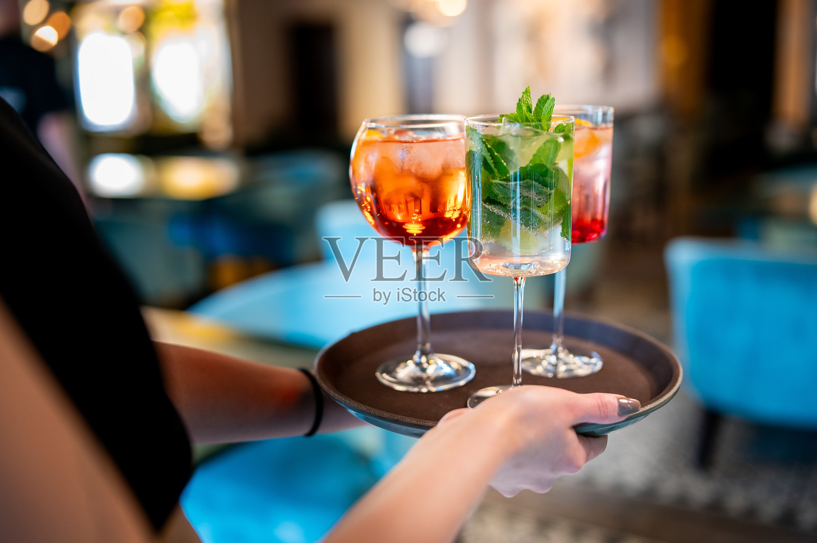 一只手拿着一个托盘，上面放着两杯装饰有薄荷叶的粉色鸡尾酒。背景模糊，暗示着高档酒吧或餐厅的环境。非常适合款待和社交活动的主题。照片摄影图片