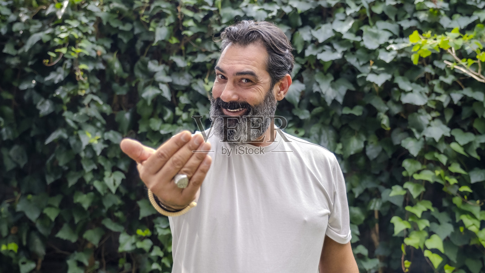 一位中年、肤色较白、留着胡子的男子，皮肤较暗，在树林中近距离拍摄，伸出手做出“过来”的手势，正看着镜头。照片摄影图片