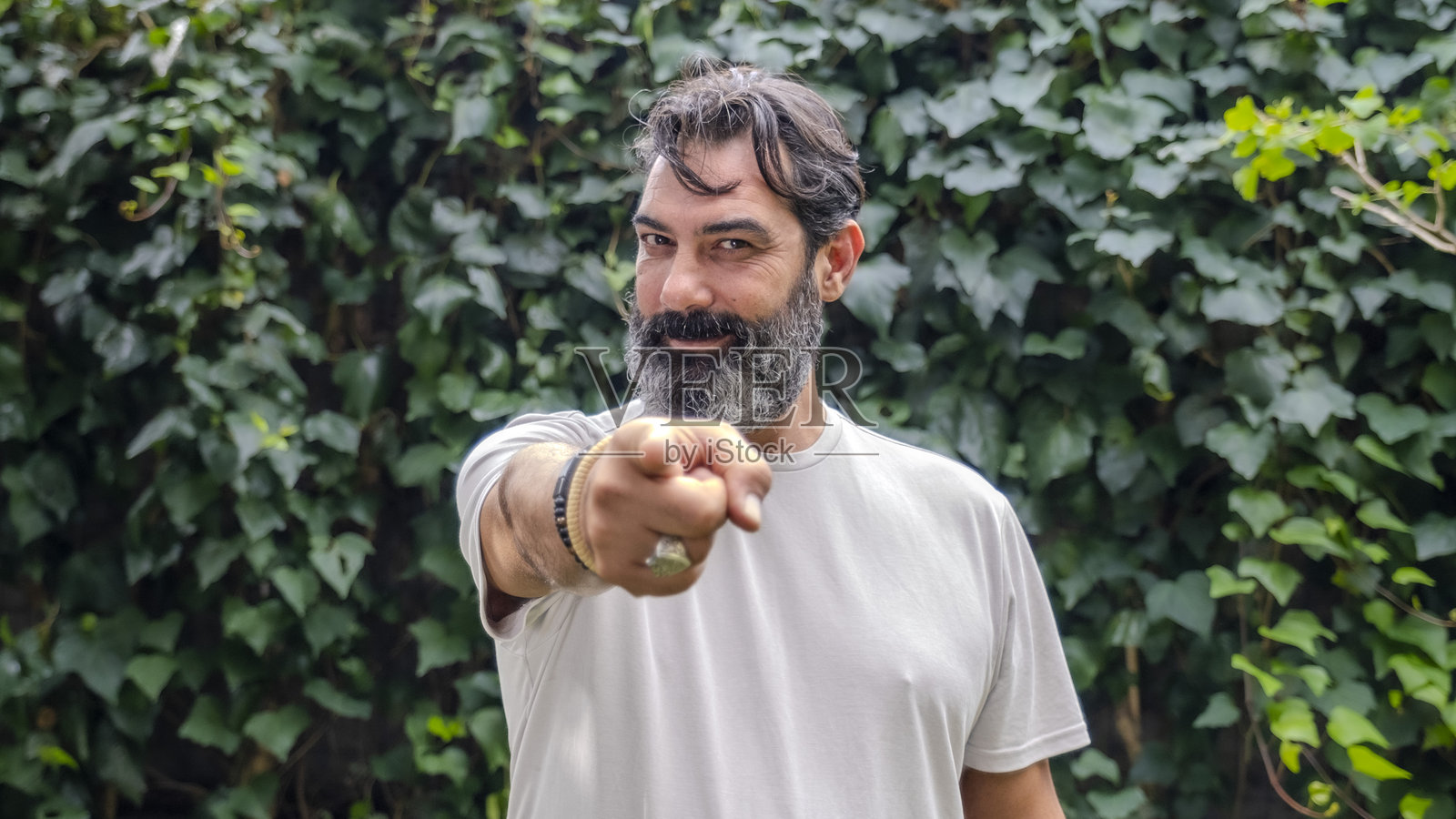 一位中年、肤色白皙、留着胡须、黑发的男子在树林的户外环境中，正对着镜头用食指指向镜头。照片摄影图片