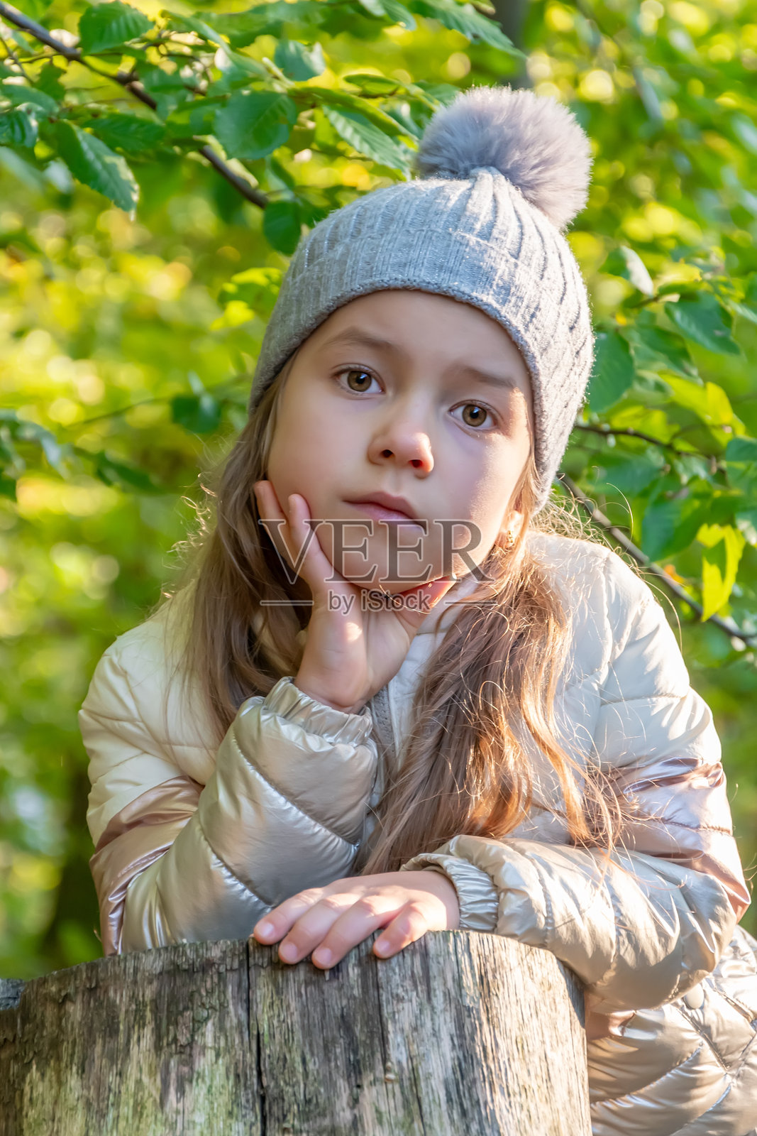 一个小女孩靠在密林中的木桩上，戴着灰色帽子和浅色夹克，展现出顽皮而又沉思的神情。照片摄影图片