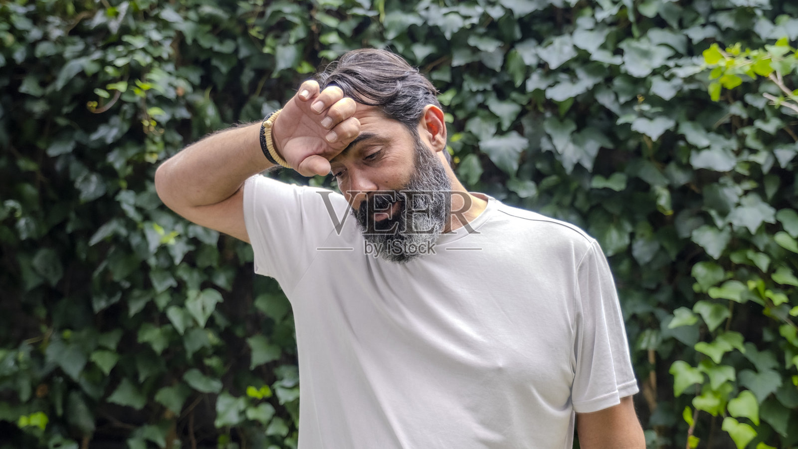 一位中年、深色头发的男子，留着胡须，肤色较白，身处树林中的户外，特写镜头中面向镜头，表情显露出因炎热而感到不适。照片摄影图片