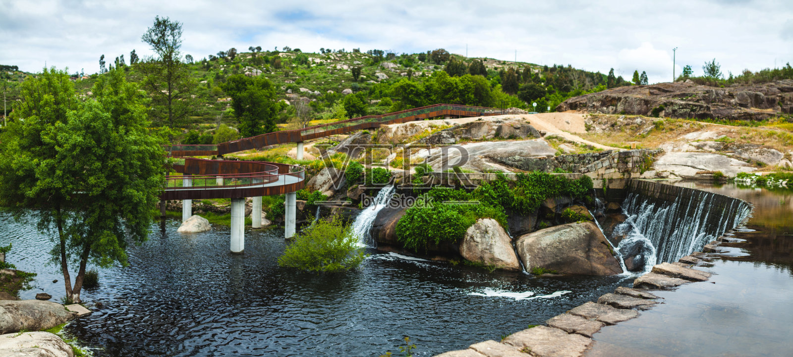 现代人行道蜿蜒在宁静的河流上方，附近有一处小瀑布，周围是绿色的山丘和岩石地形，天空多云。照片摄影图片