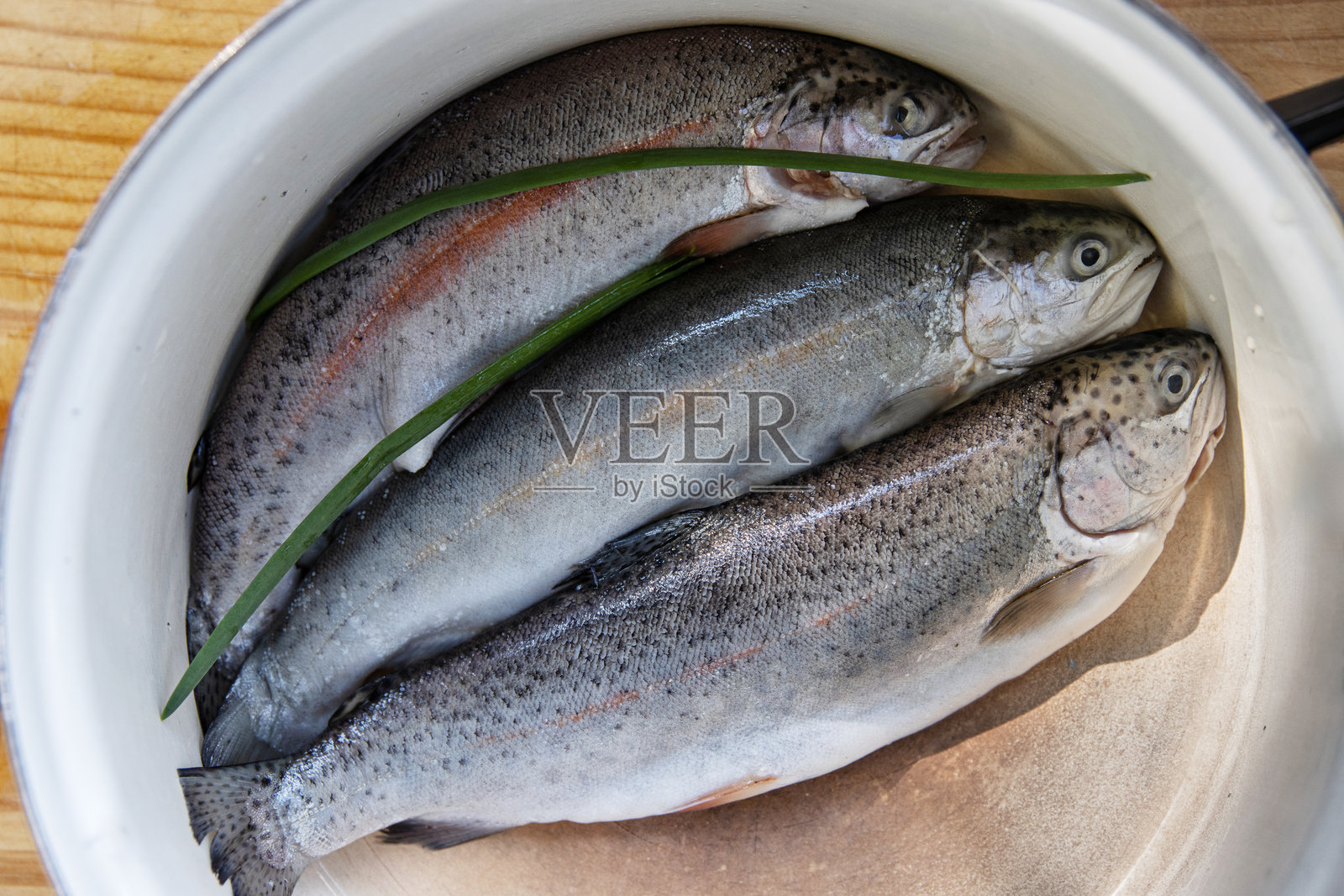 三条新鲜捕获的彩虹鳟鱼放在厨房的白色搪瓷碗里。特写镜头展示了一条新鲜的鳟鱼在炖锅中，准备进行烹饪。俯视图。照片摄影图片