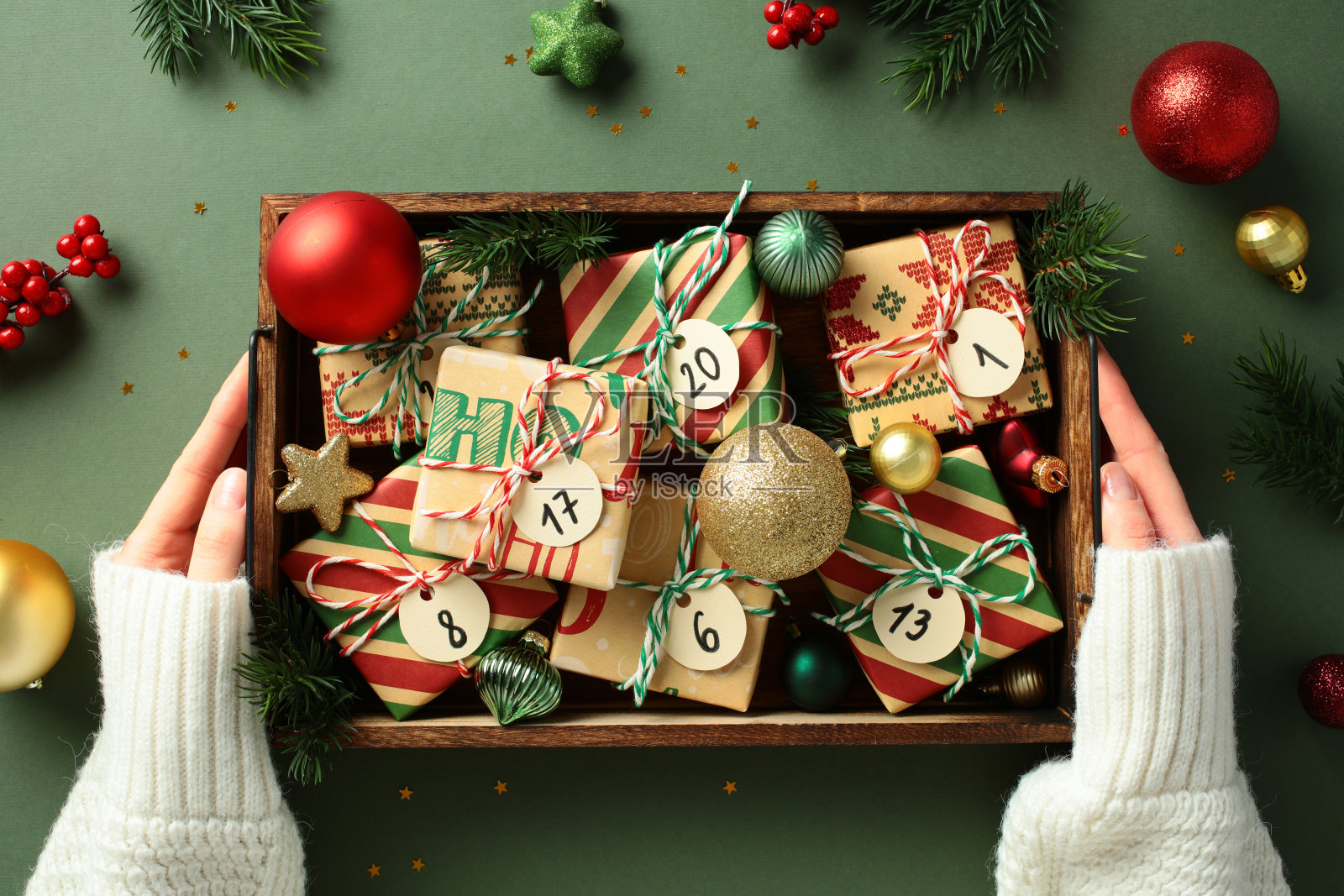 女性手中拿着装有编号的圣诞日历标签的木盒，背景为绿色，周围有节日装饰品和松枝。照片摄影图片