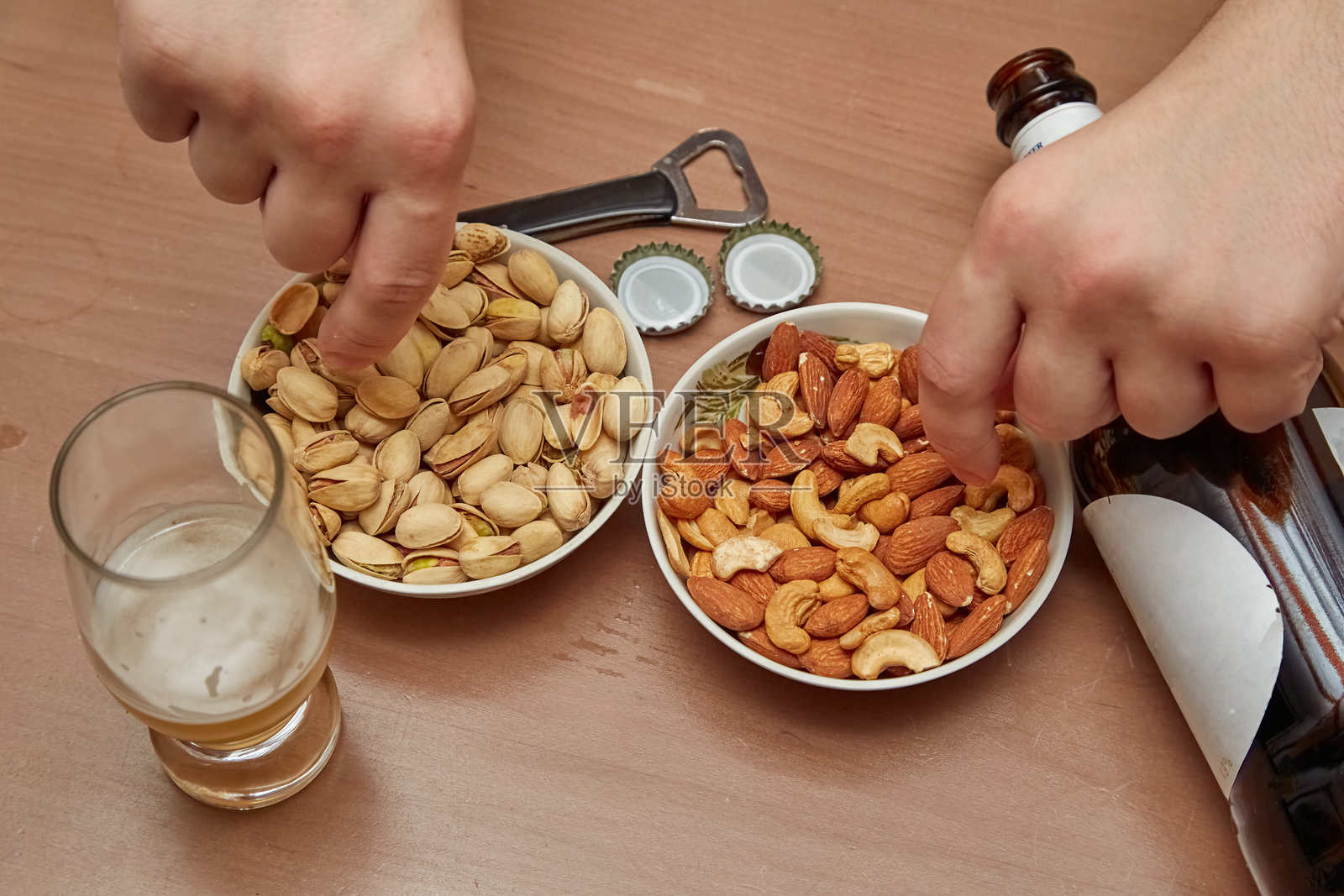 两只手从盘子里拿坚果，桌上有开心果和坚果，还有一瓶啤酒、一杯啤酒和一个开瓶器，啤酒小吃，酒吧小吃。照片摄影图片