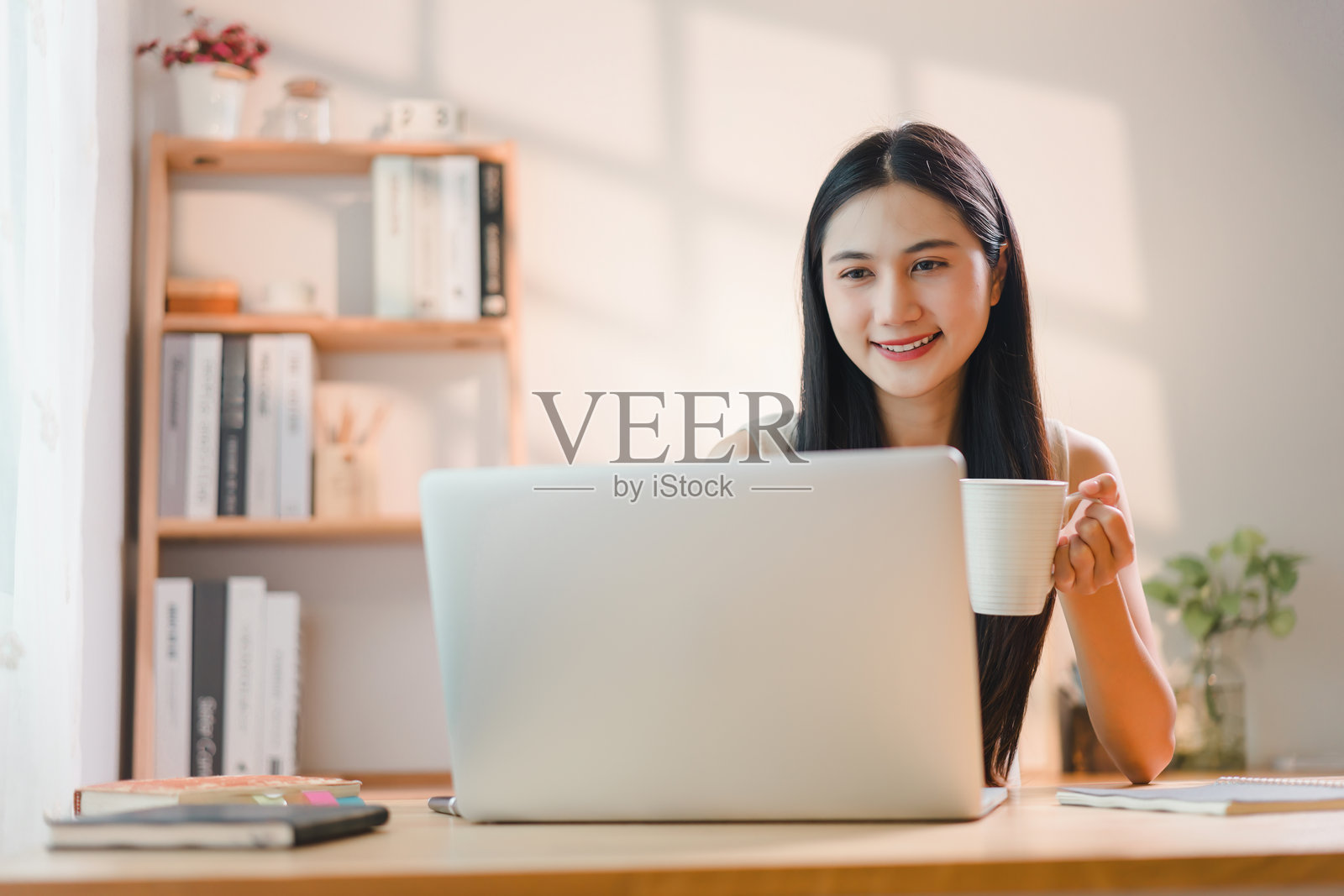 年轻的亚洲女性在家中办公室使用笔记本电脑，同时享用饮品。照片摄影图片