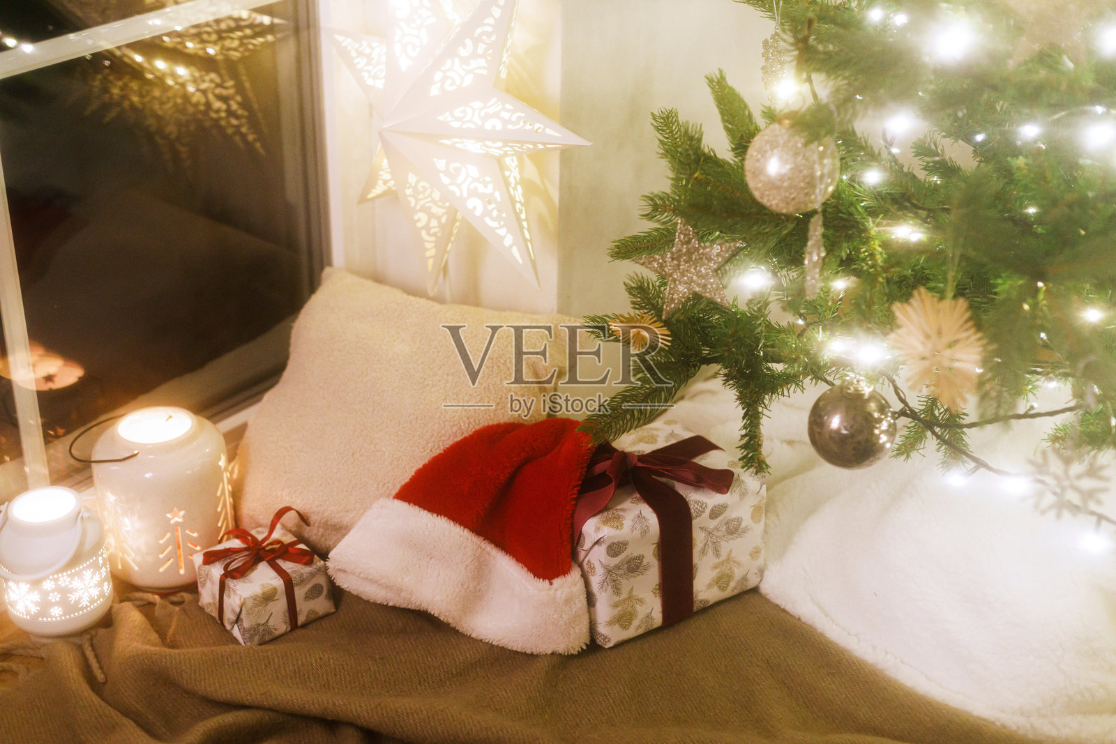 圣诞快乐！时尚的圣诞礼物、发光的灯笼、柔软舒适的枕头和圣诞老人帽子在灯光下的圣诞树下。温馨的圣诞夜氛围，柔和的冬季舒适静物。照片摄影图片