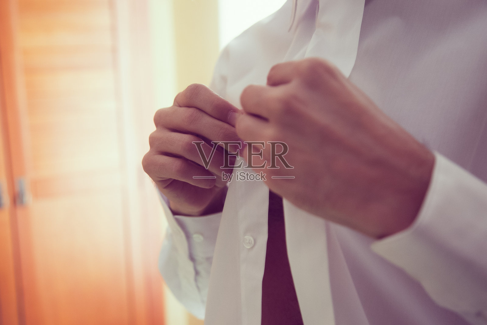 一名男子在光线明亮的房间里扣上他的白衬衫，背景是木制橱柜。照片摄影图片