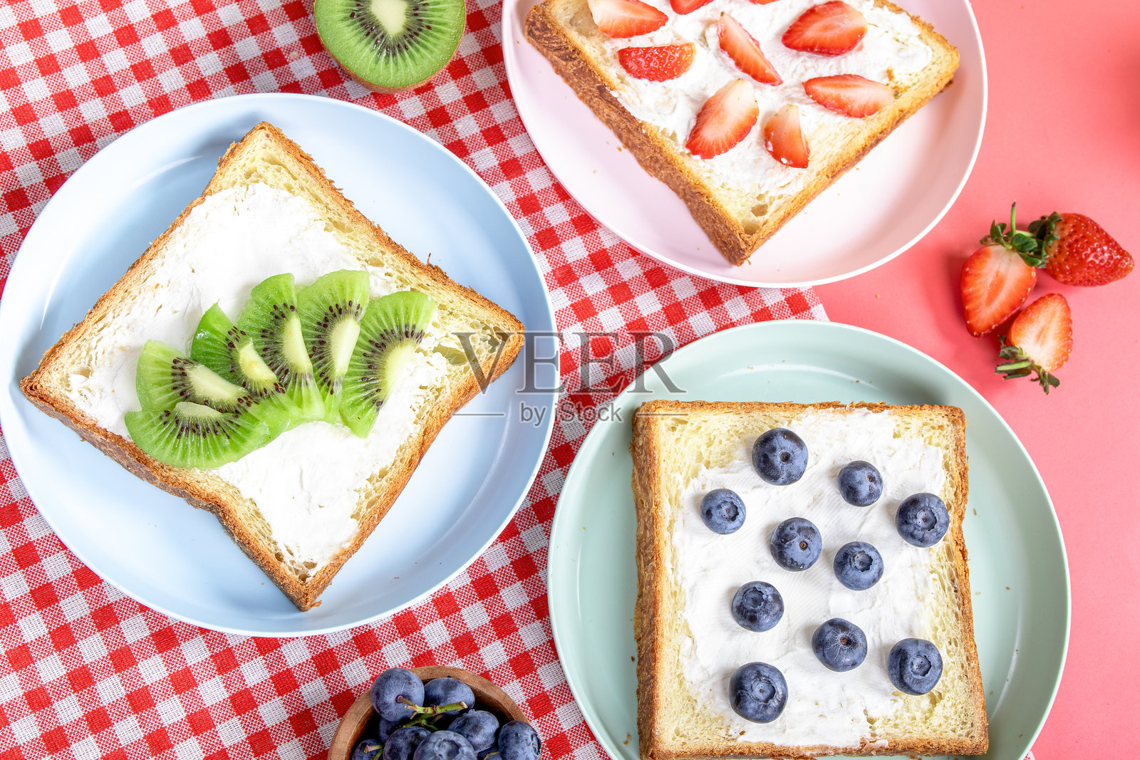 各种水果吐司，上面放有蓝莓、草莓和猕猴桃，搭配白面包，摆放在五彩斑斓的盘子上，背景是红色格子布，旁边有切片面包、整颗水果和书籍。照片摄影图片
