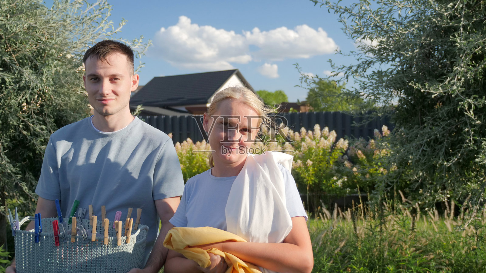 年轻男子和中年女性微笑着在花园里手持洗衣用品，营造出阳光明媚的温馨家庭氛围。照片摄影图片