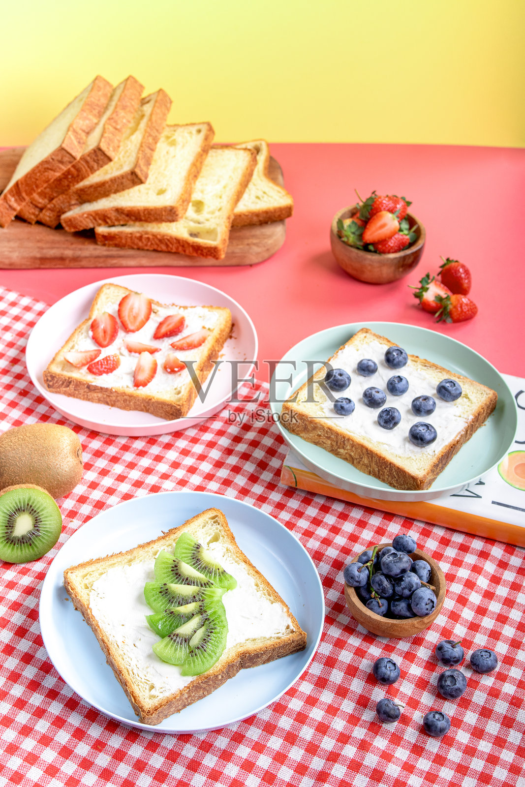 各种水果吐司，上面放有蓝莓、草莓和猕猴桃，搭配白面包，摆放在五彩斑斓的盘子上，背景是红色格子布，旁边有切片面包、整颗水果和书籍。照片摄影图片