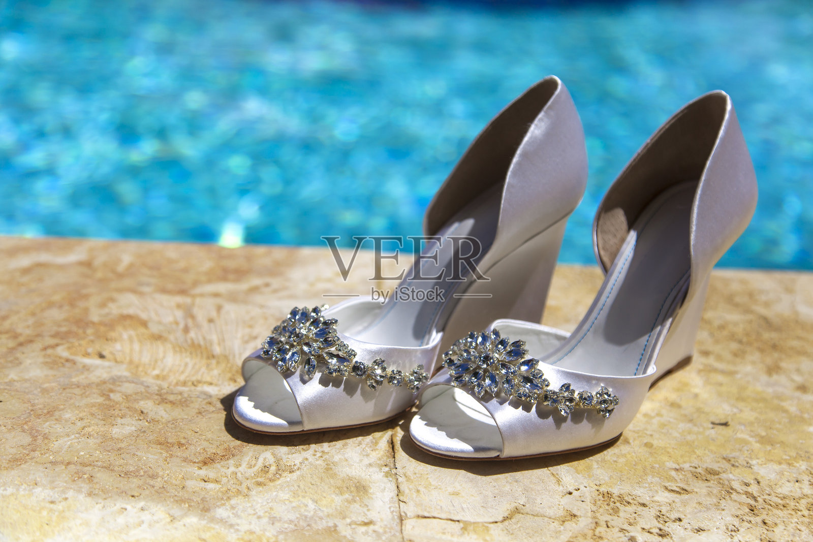 优雅的白色高跟鞋，饰有装饰物，放置在闪闪发光的蓝色泳池旁边照片摄影图片