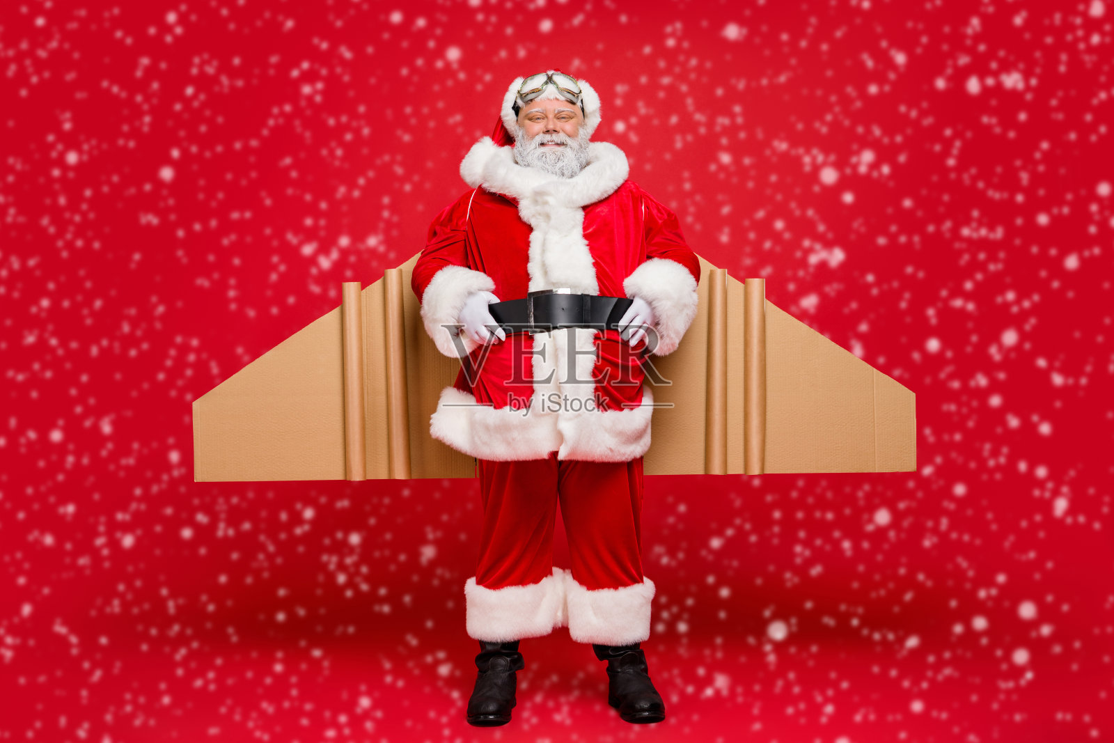 全身照的老年时尚圣诞老人，戴着帽子，拥有纸板手工翅膀，准备在圣诞夜飞翔，穿着魔法服装和护目镜，背景为红色。照片摄影图片