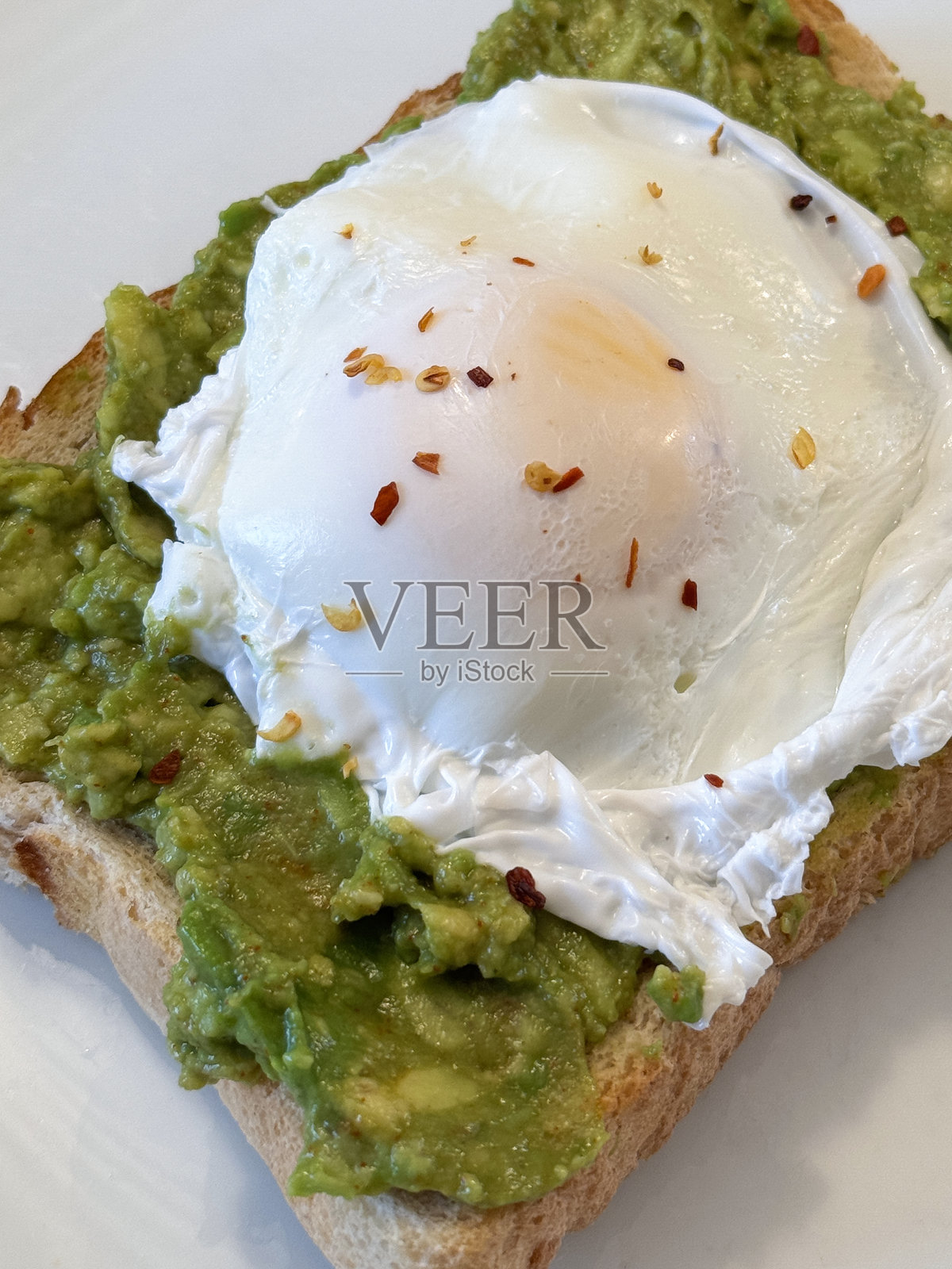 特写镜头，白色盘子里盛着早餐：白面包吐司，上面放着捣碎的鳄梨和水煮蛋，撒上干红辣椒碎，俯视角度，重点在前景。照片摄影图片