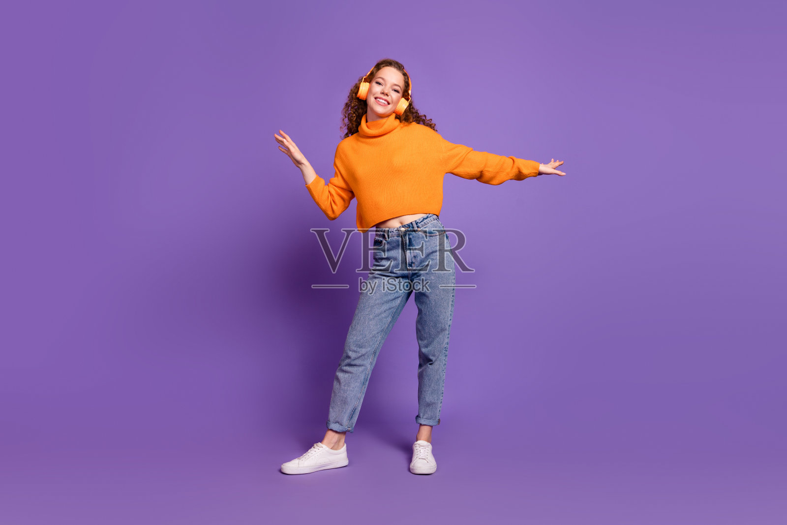 全身照，漂亮的年轻女孩戴着耳机跳舞，穿着毛衣，背景为紫色。照片摄影图片