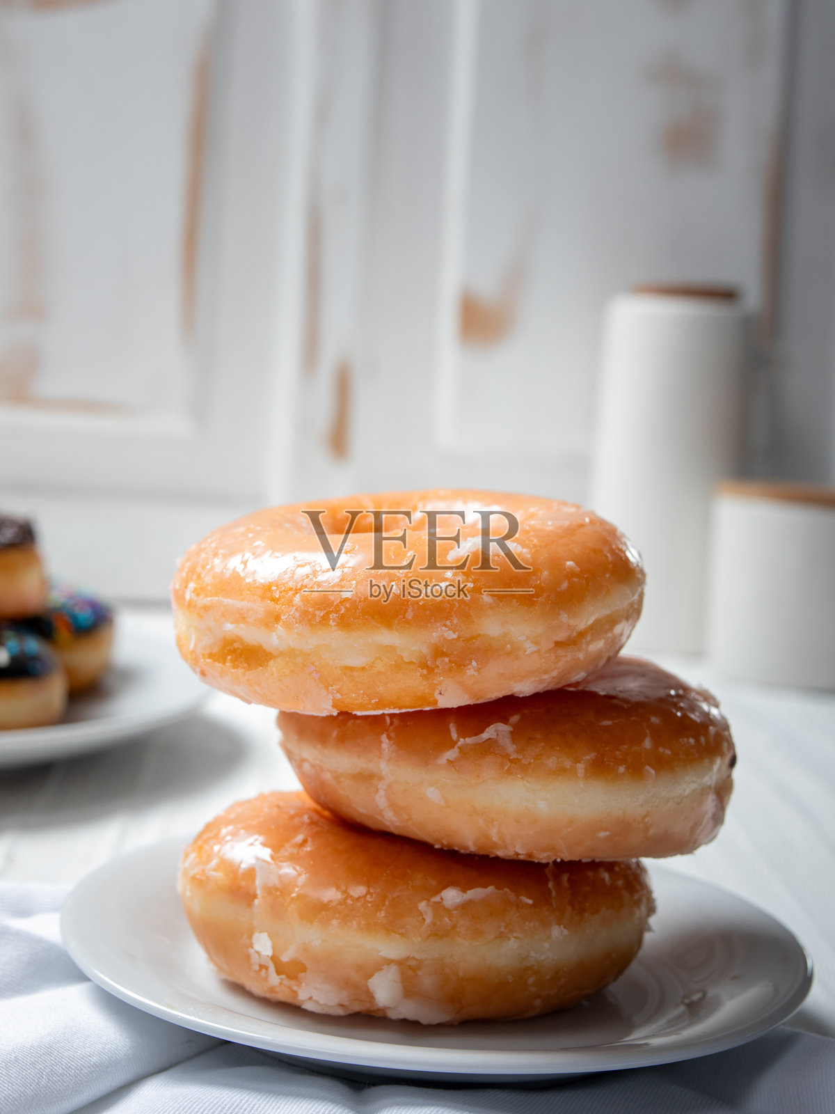 一盘放在白色盘子上的釉面甜甜圈。背景中还有另一盘各种甜甜圈，其中包括一个撒有巧克力糖屑的甜甜圈。场景设定在一个明亮通透的厨房里。照片摄影图片