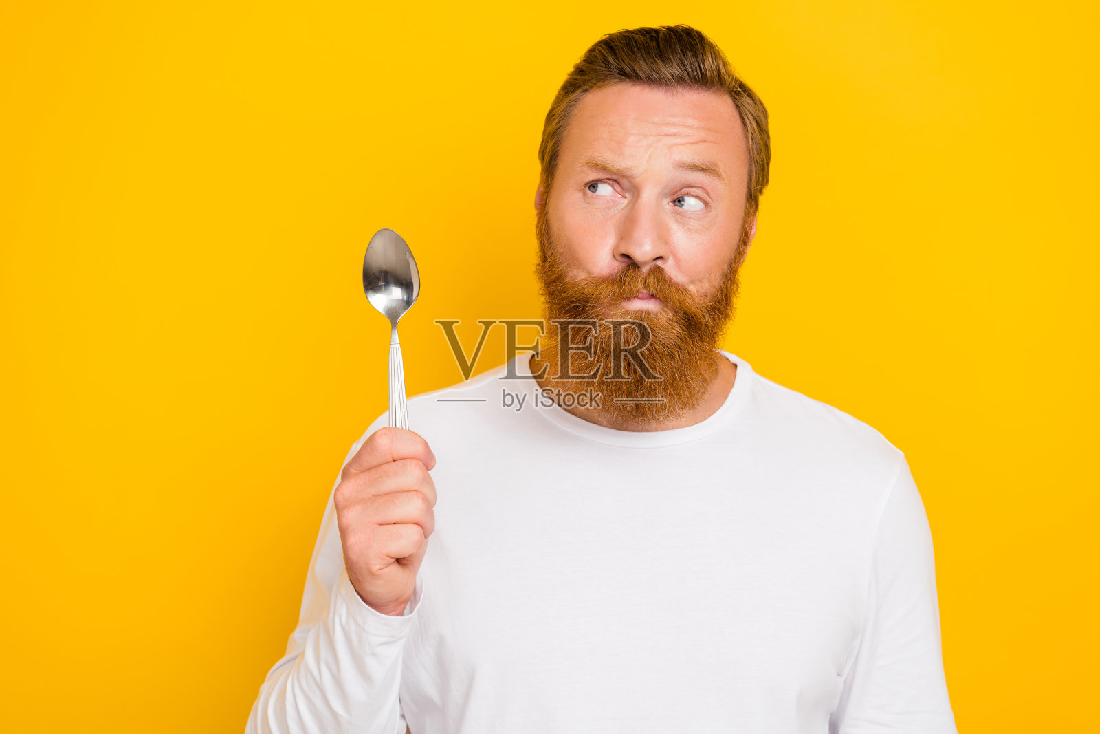 年轻男子的照片肖像，怀疑的表情，手持勺子，穿着时尚的白色服装，背景为黄色。照片摄影图片