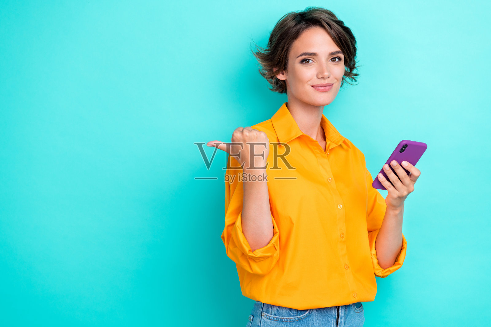 漂亮女士手持电话，手臂指向空旷空间，展示现代设备，出售酷炫产品，背景为青色。照片摄影图片