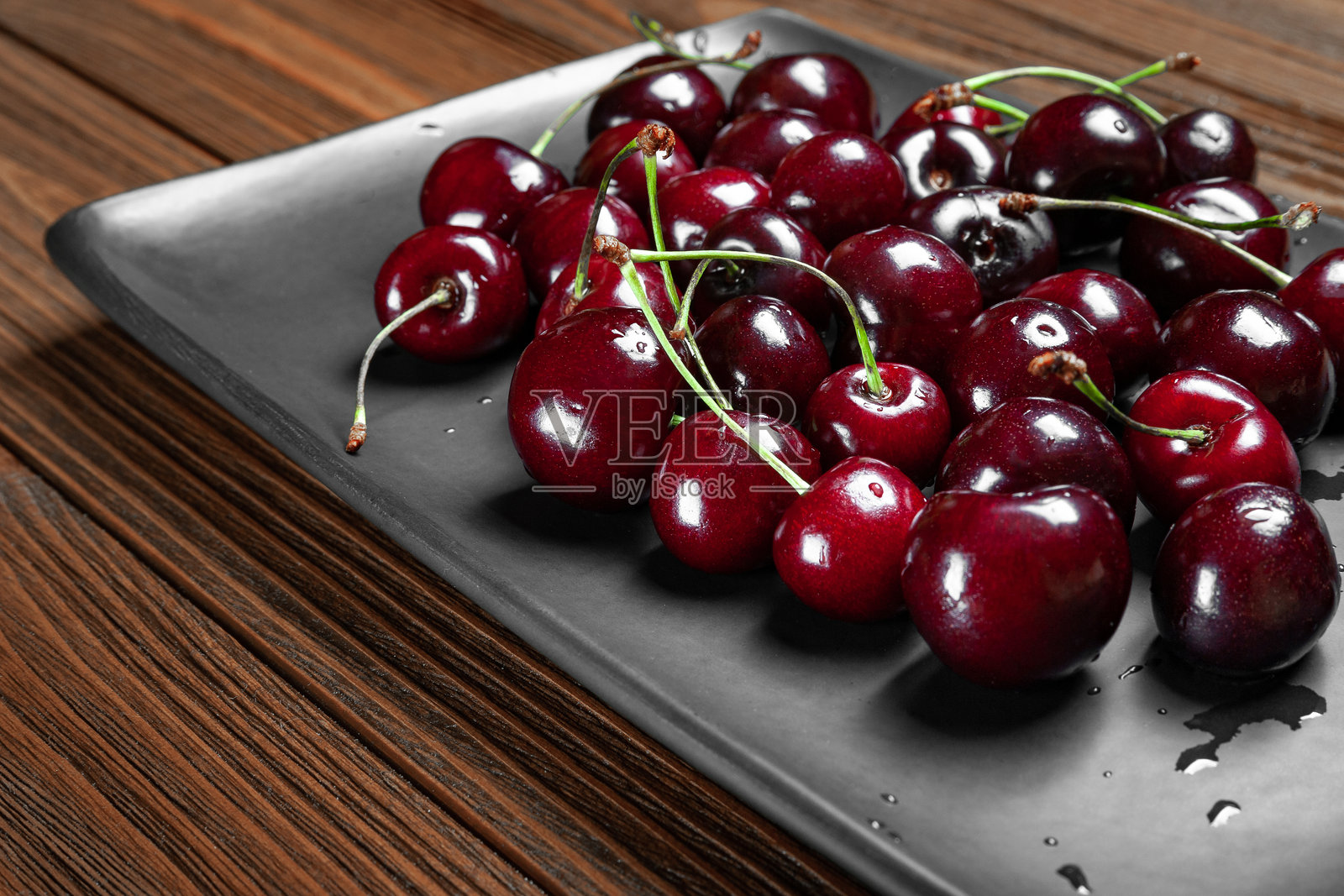 新鲜成熟多汁甜美的樱桃放在黑色盘子里，背景是木质的。湿润的新鲜樱桃上有水珠。照片摄影图片