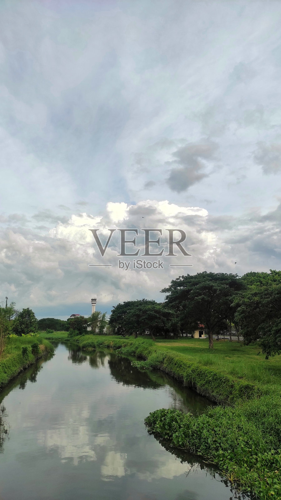 宁静的河边景色，郁郁葱葱的绿树在阴云密布的天空下。平静的河水映照着周围的树木，远处高耸的塔楼为这幅宁静的风景增添了深度。照片摄影图片