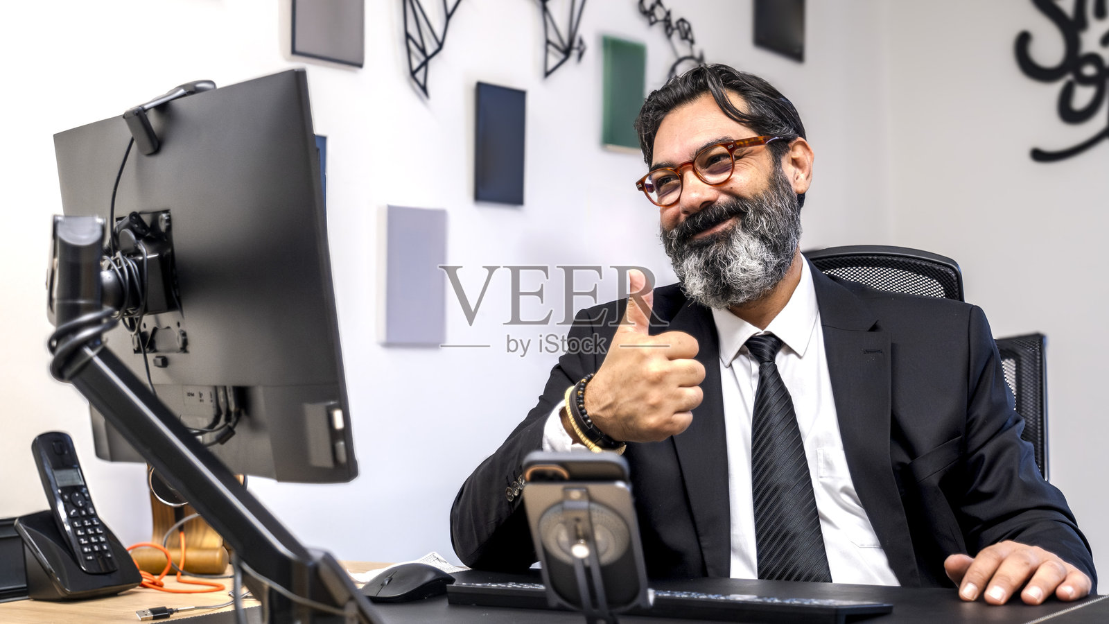 一位中年、深色头发、留着胡子的商人在办公室的桌子前，正在电脑上进行视频会议，竖起大拇指祝贺他的员工们。照片摄影图片