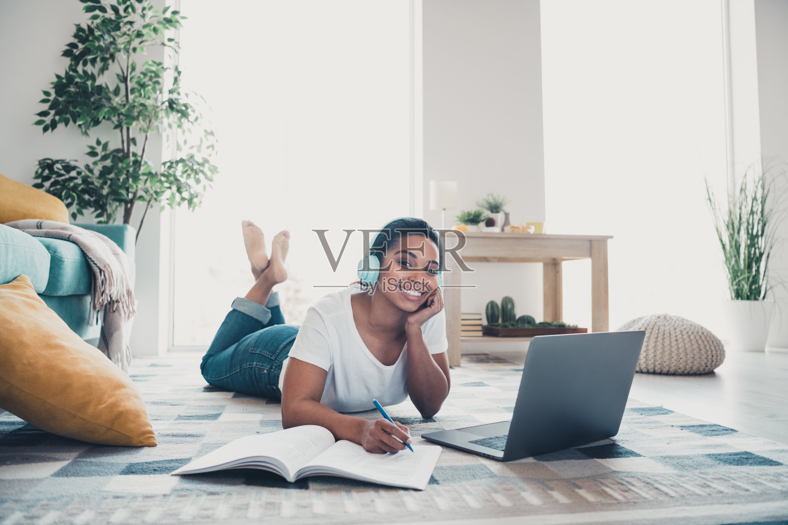 全身肖像，年轻女性佩戴耳机，使用笔记本电脑，手持钢笔，穿着衬衫，现代室内公寓环境。照片摄影图片