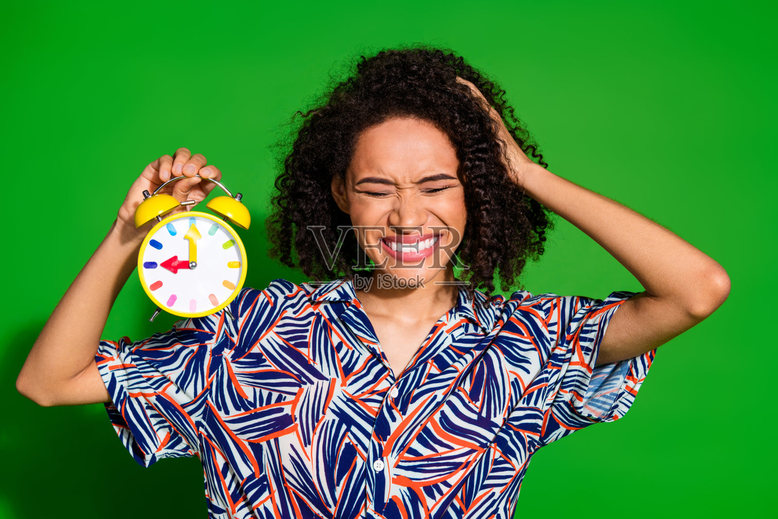 一位迷人的年轻女性手持时钟，表情恼怒，穿着时尚多彩的衣服，背景为绿色。照片摄影图片