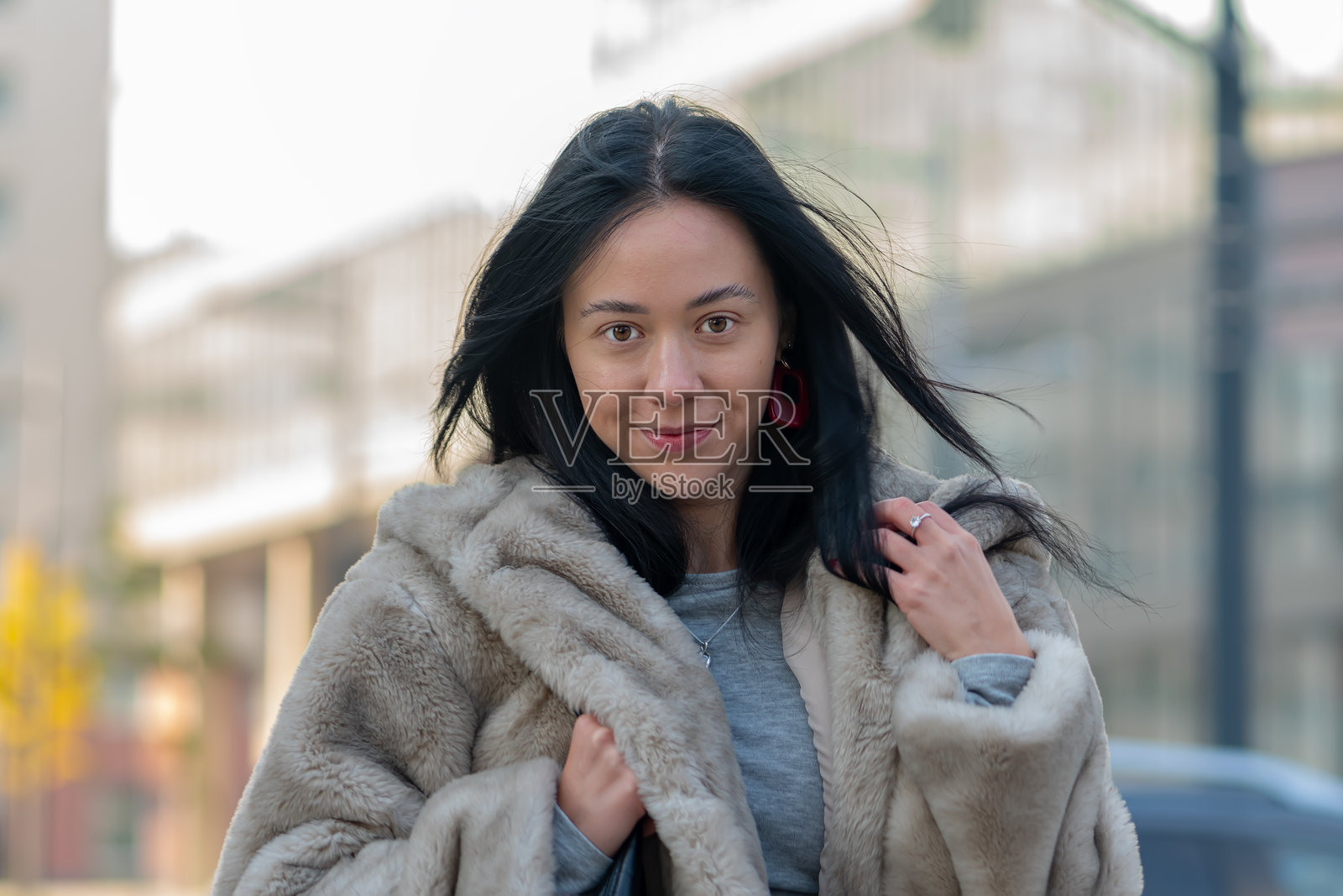 一位长发黑色、具有东方特征的女性穿着温暖的毛皮外套，站在阳光明媚的城市街道上。照片摄影图片