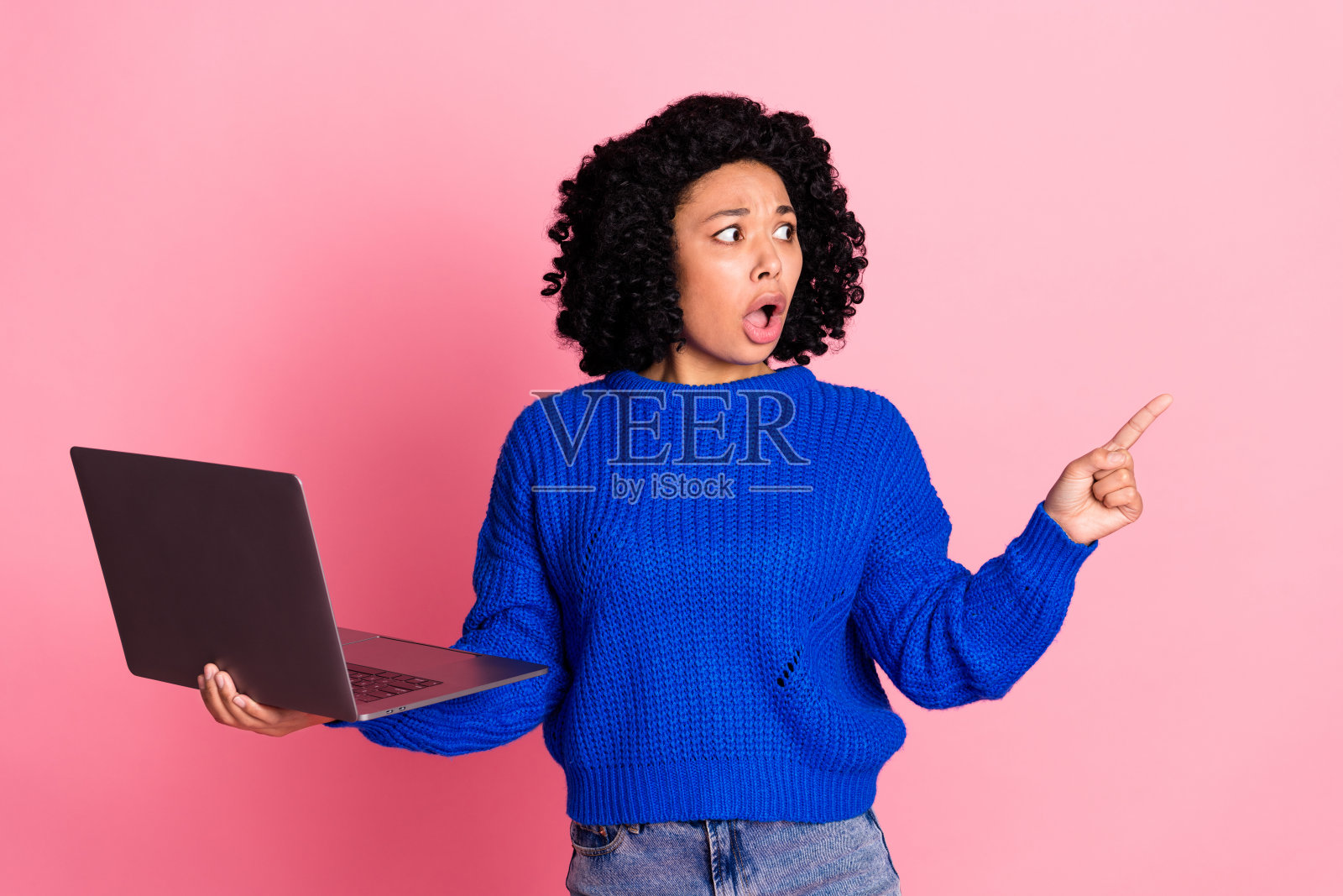 一位黑色波浪卷发的女士穿着蓝色毛衣，使用笔记本电脑，手指指向令人震惊的价格，背景为粉色。照片摄影图片