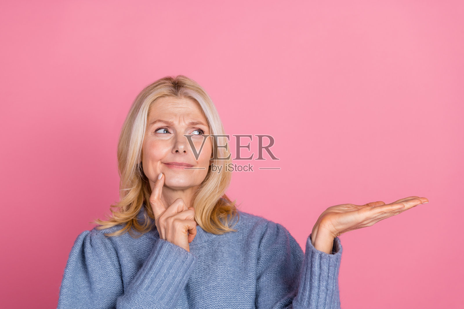 穿着时尚蓝色针织衣服的迷人老年女性，面露不确定的神情，目光投向空旷的空间，背景为粉色。照片摄影图片