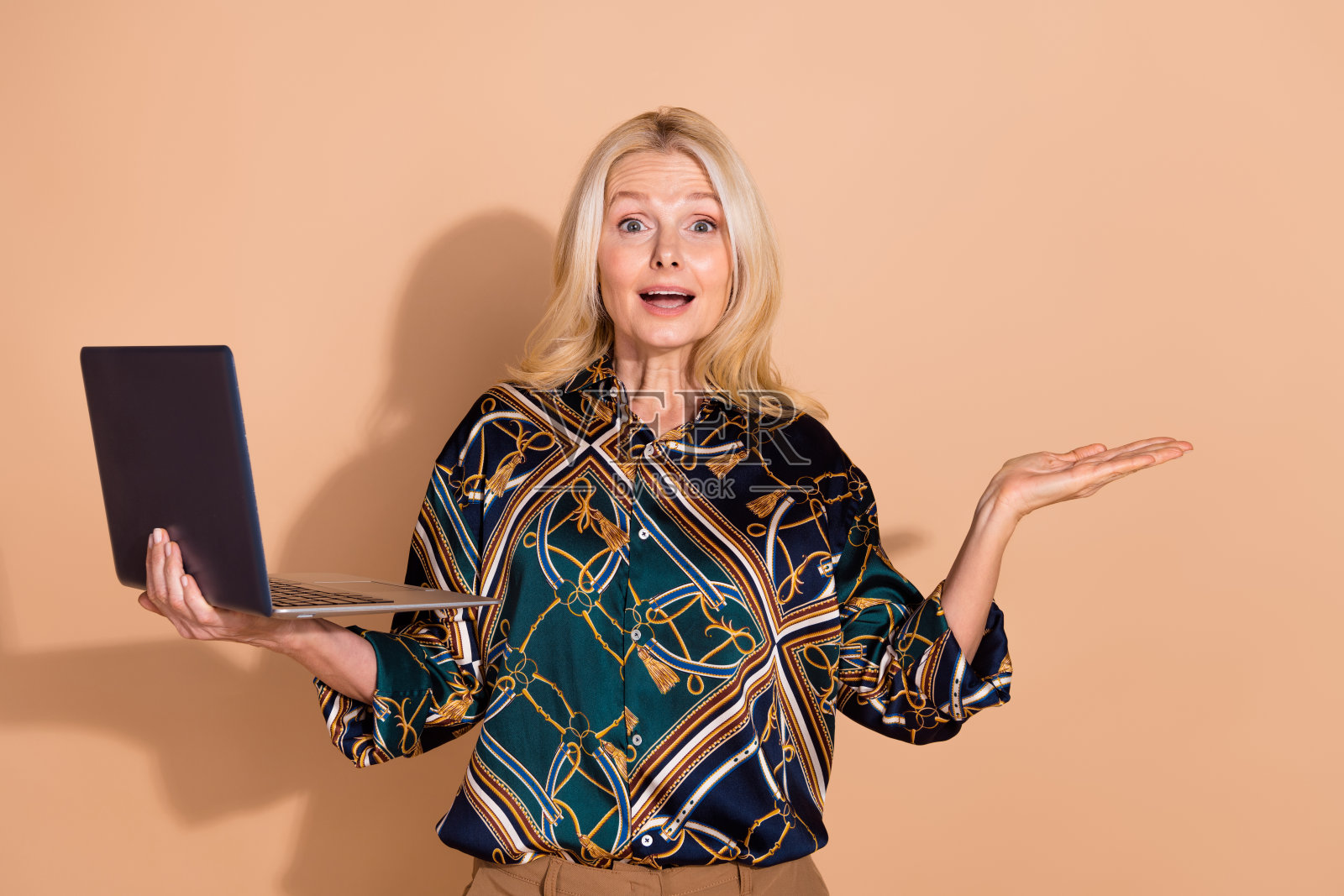 金发女商人在优雅衬衫中使用笔记本电脑，手中拿着促销Facebook广告，背景为米色。照片摄影图片