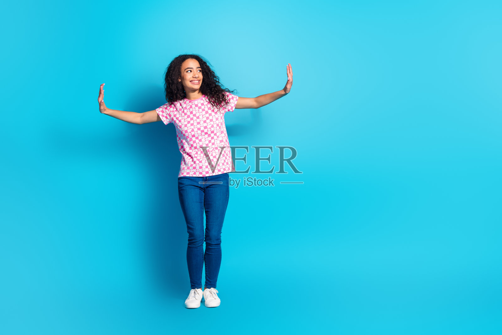 全身照，迷人积极的女孩穿着印花T恤，双臂向空旷的空间伸展，背景为蓝色。照片摄影图片