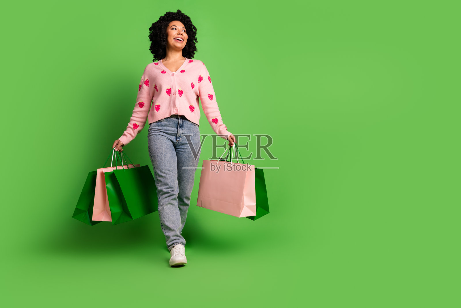 全身照，漂亮女孩手持购物袋，望向空旷的地方，穿着草莓印花的粉色服装，背景为绿色。照片摄影图片