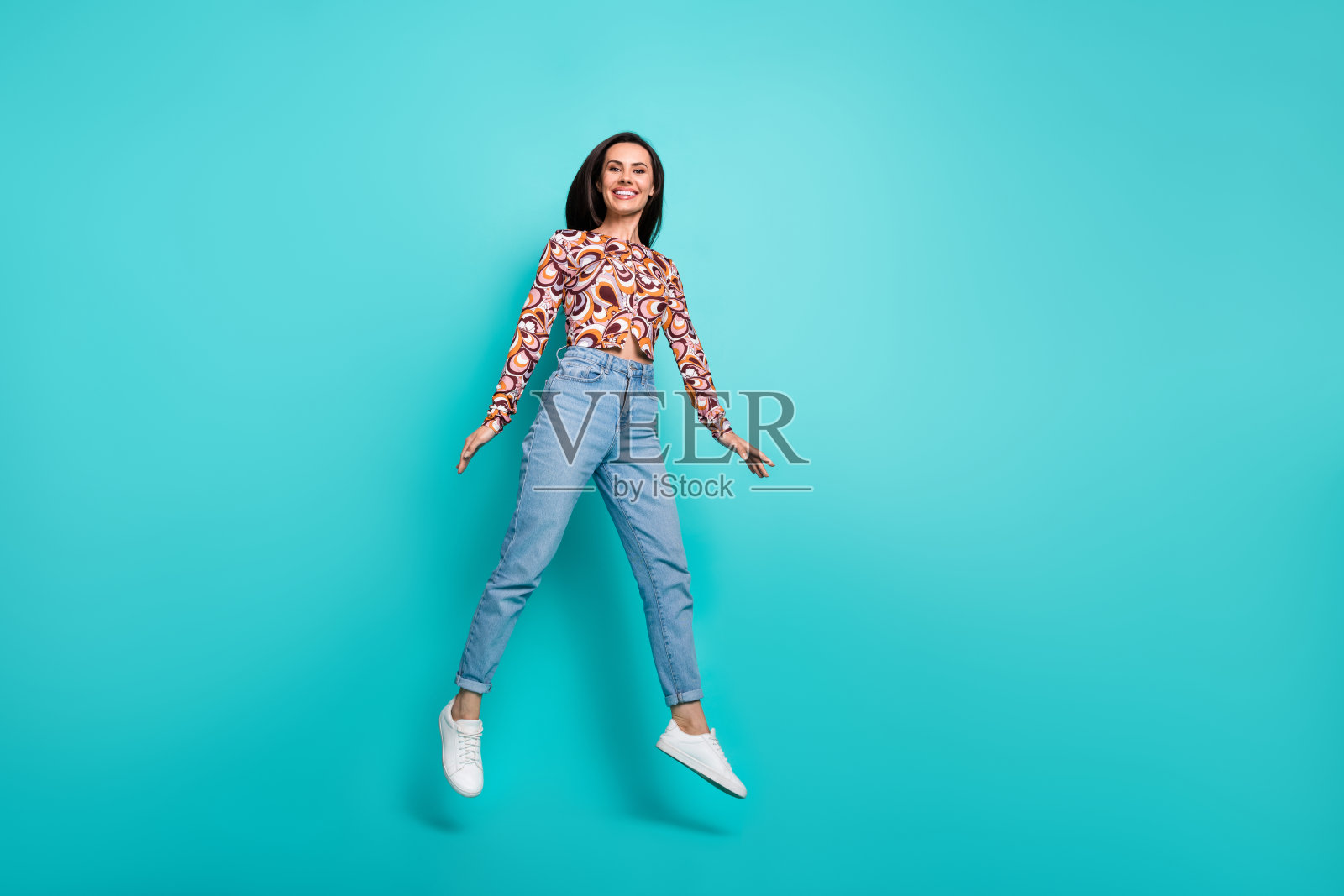 充满魅力的积极女性穿着印花衬衫在空旷的空间中高高跳起，背景为青绿色。照片摄影图片