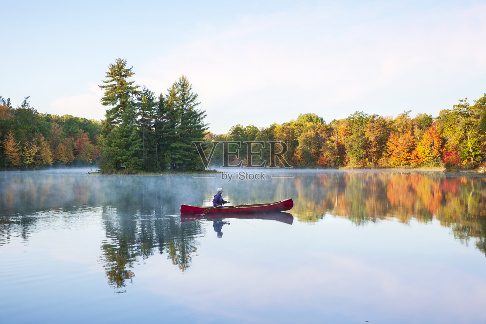 一位老年男子在美丽的明尼苏达州北部湖泊上划着一只红色木制独木舟，清晨的秋天，湖中有一个小岛。照片摄影图片