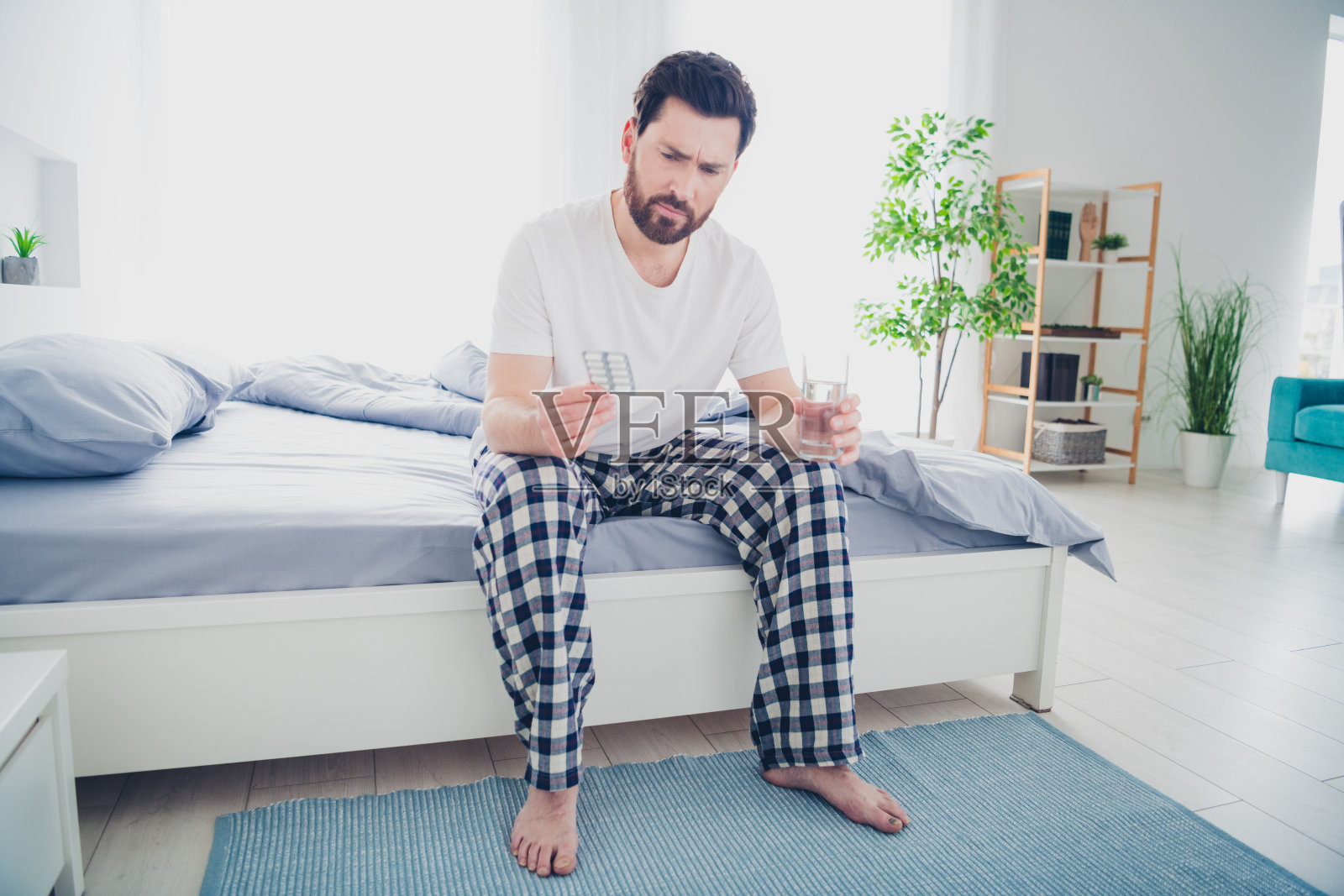 穿着睡衣的年轻男子坐在床上，神情紧张和怀疑，正在室内公寓里喝药。照片摄影图片