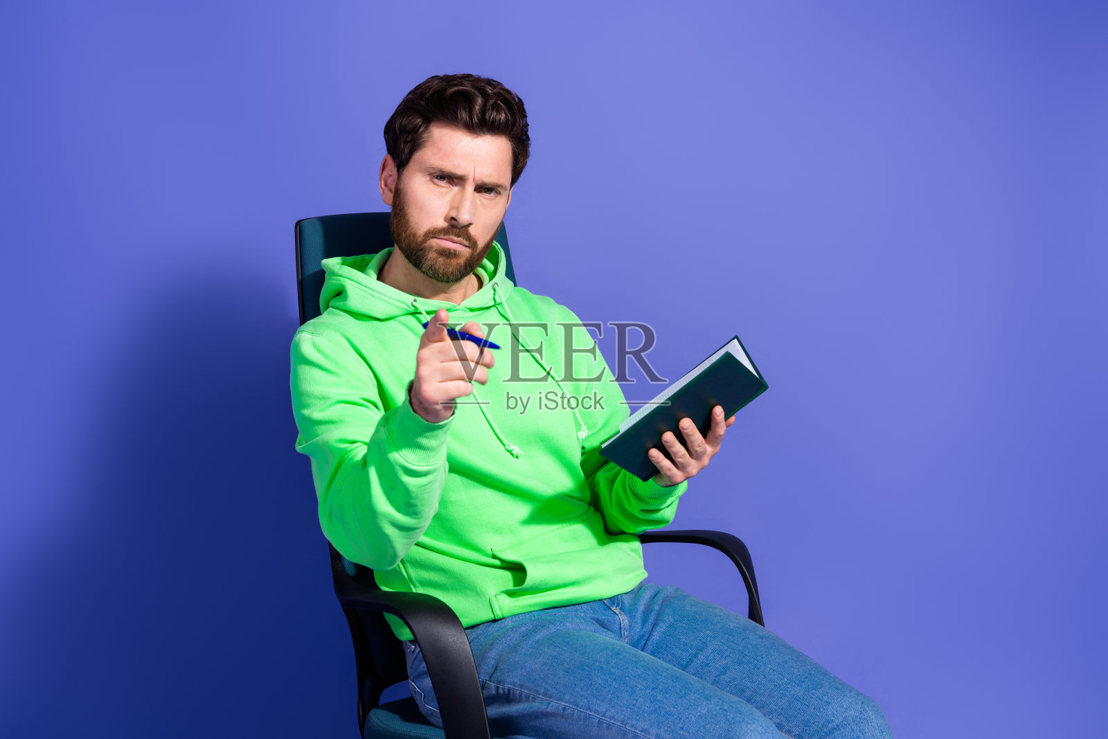 穿着时尚绿色服装的年轻帅气男性坐在扶手椅上，认真地指着你，手里写着笔记本，背景为紫色。照片摄影图片