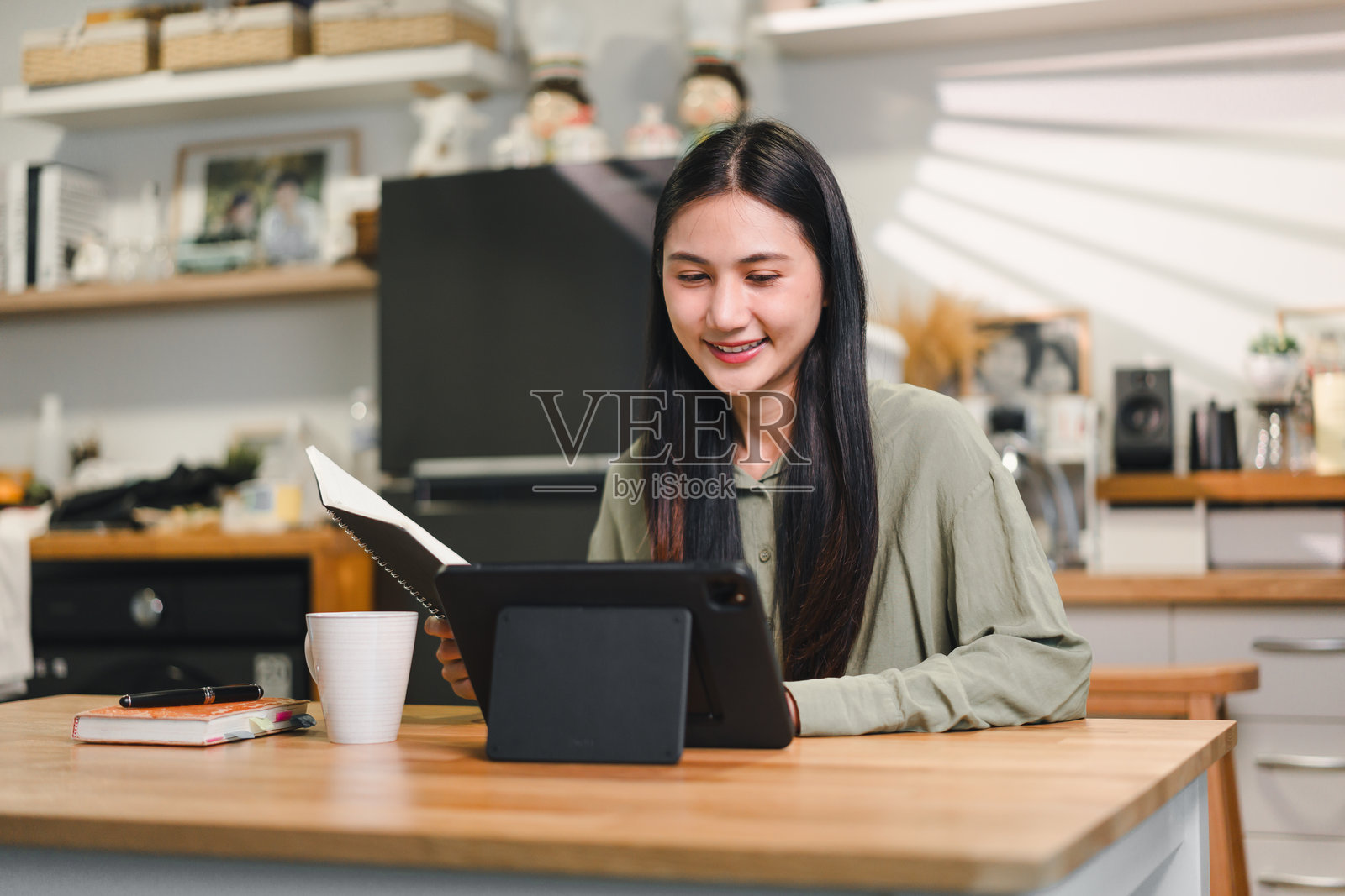 年轻女性坐在厨房桌子旁，微笑着使用平板电脑并在笔记本上写字。温馨的厨房环境具有现代装饰和温暖的灯光，营造出一种舒适的氛围。照片摄影图片