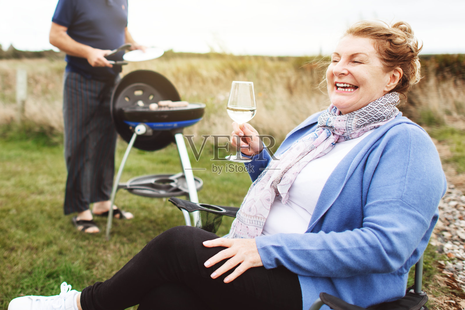 快乐的白人女性在户外享受一杯葡萄酒。女性微笑着，手持葡萄酒，放松身心。背景中有一名男性在烧烤。女性与葡萄酒，户外放松。家庭露营旅行中的烧烤。照片摄影图片