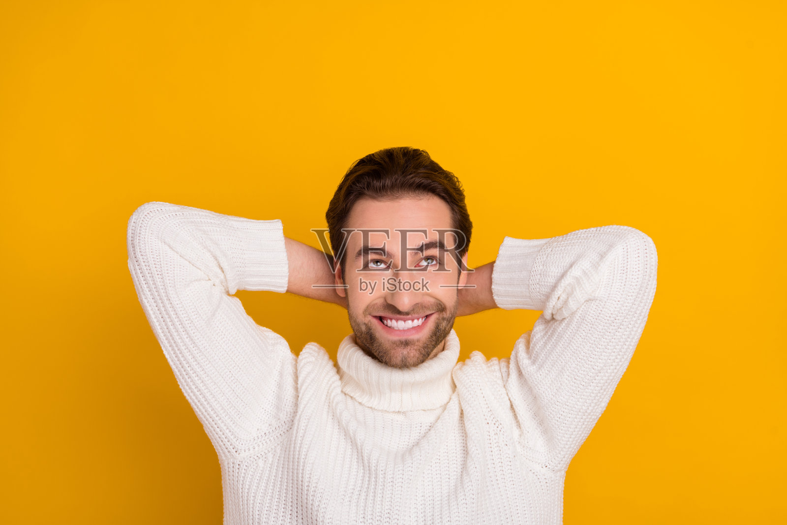 穿着白色毛衣的帅气男孩，双手放在头后，思考的姿势，背景为空旷的黄色。照片摄影图片