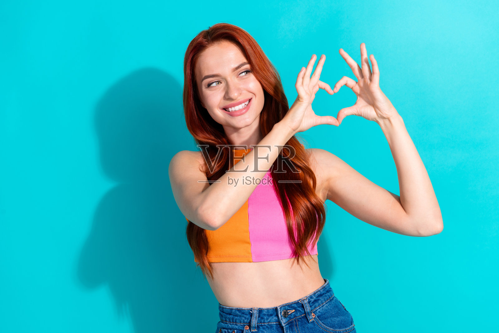 漂亮年轻女孩展示心形手势，空旷的空间，穿着上衣，背景为鲜艳的青色。照片摄影图片