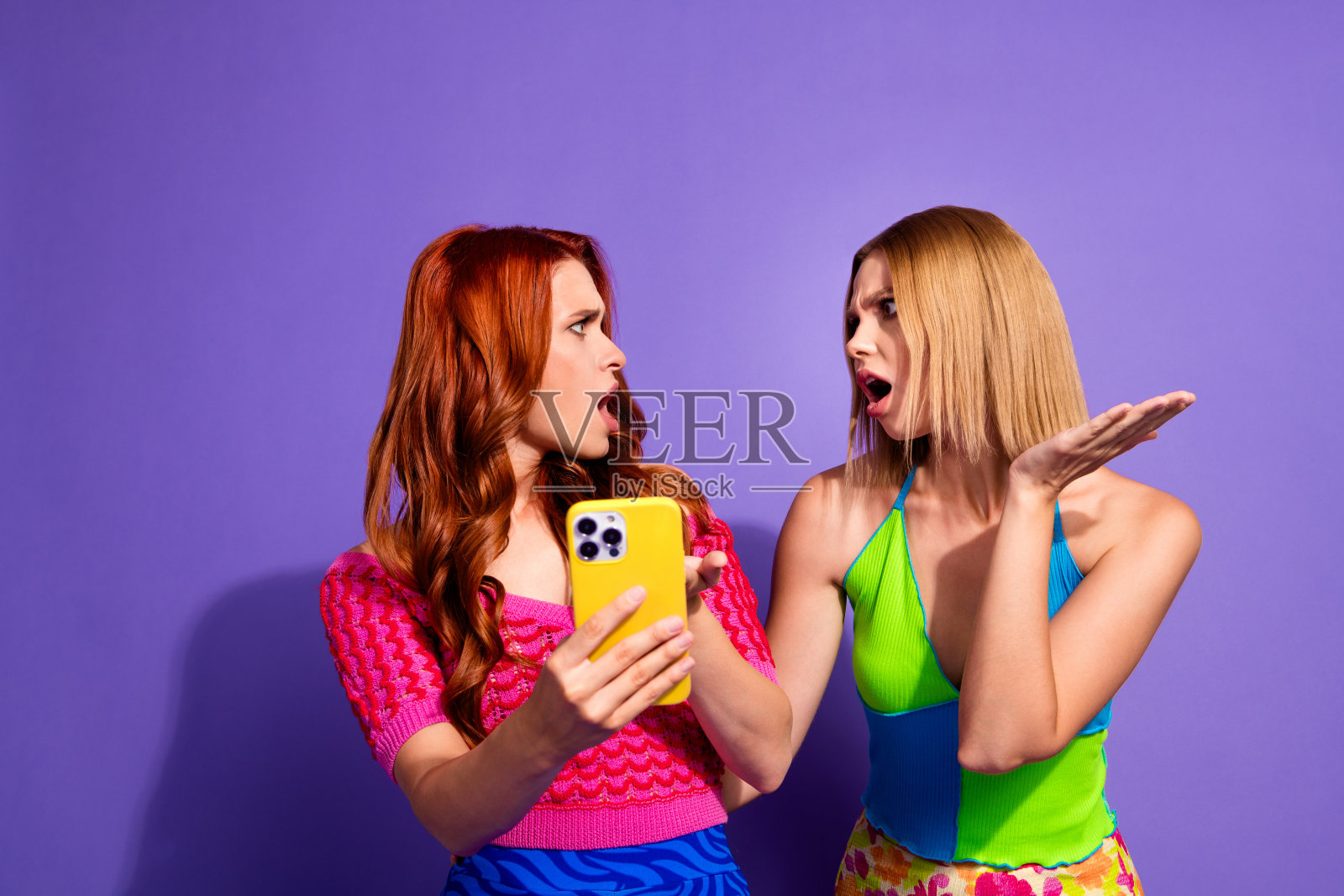 两位漂亮的女孩手持智能手机，张嘴惊讶地盯着镜头，背景为紫色。照片摄影图片