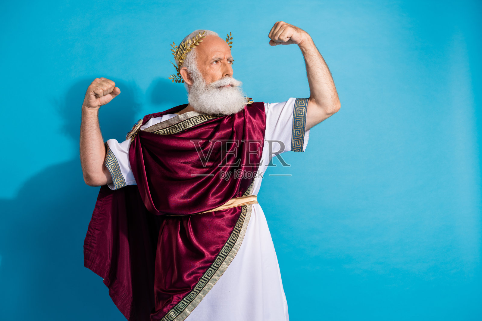 穿着希腊神帝演员服装的迷人老年男子，强壮的手臂展示出二头肌，背景为蓝色，显得空旷。照片摄影图片