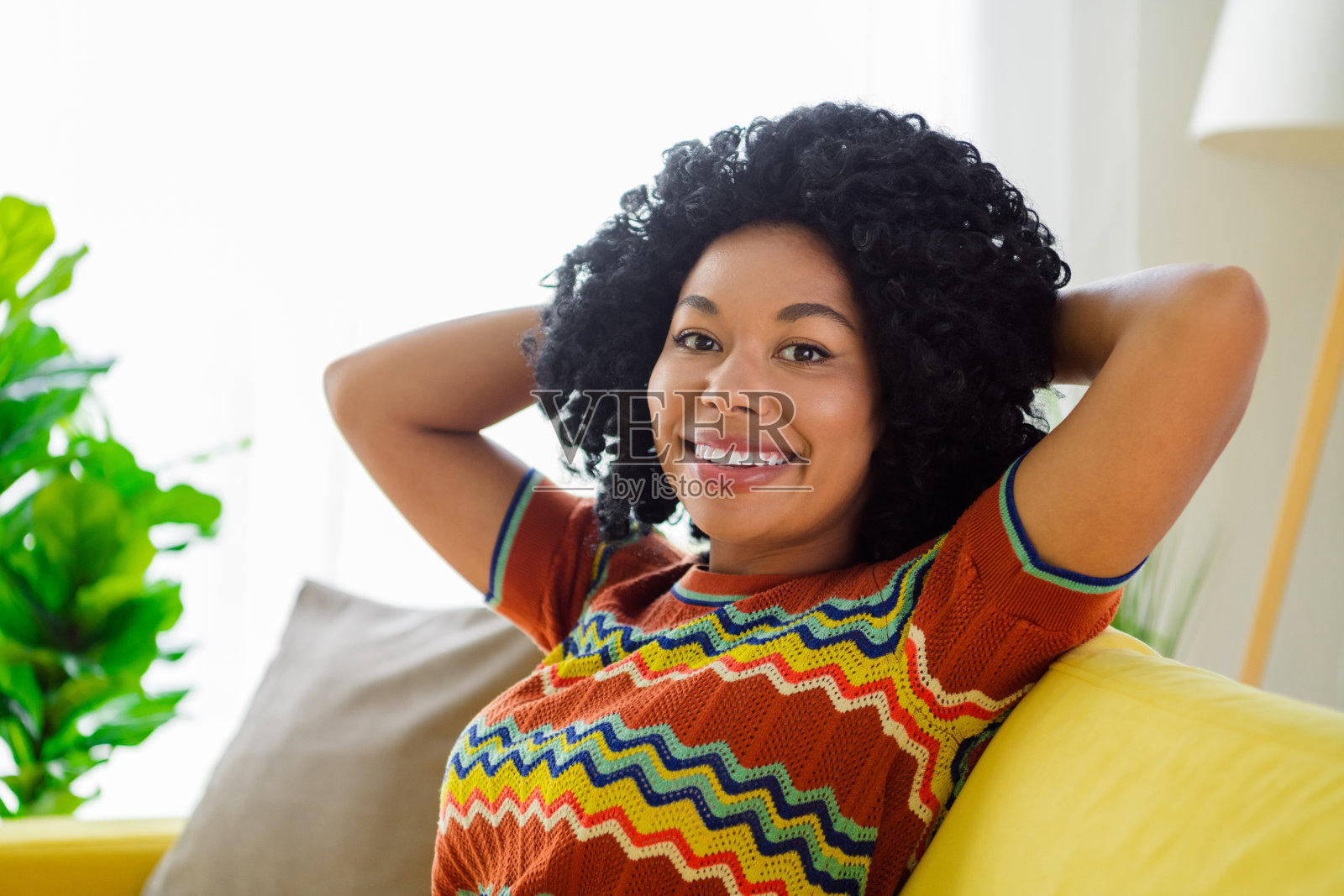 漂亮年轻女孩坐在沙发上，双臂放在头后，穿着T恤，享受现代室内公寓的闲暇时光。照片摄影图片