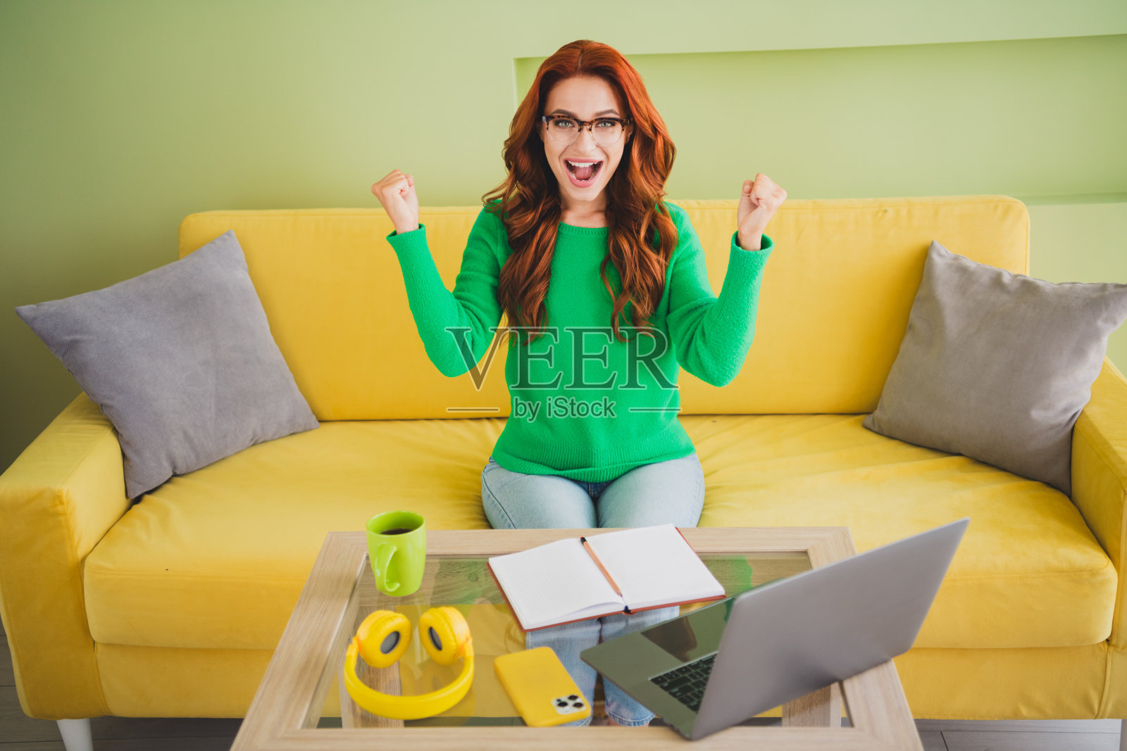 年轻女孩坐在沙发上，举起拳头，桌上放着笔记本电脑，穿着毛衣，享受周末，现代绿色室内环境。照片摄影图片