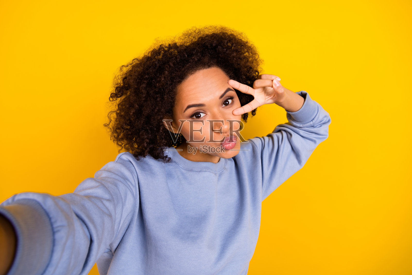 迷人卷发女孩自拍，做出V字手势，亲吻嘴唇，背景为黄色。照片摄影图片