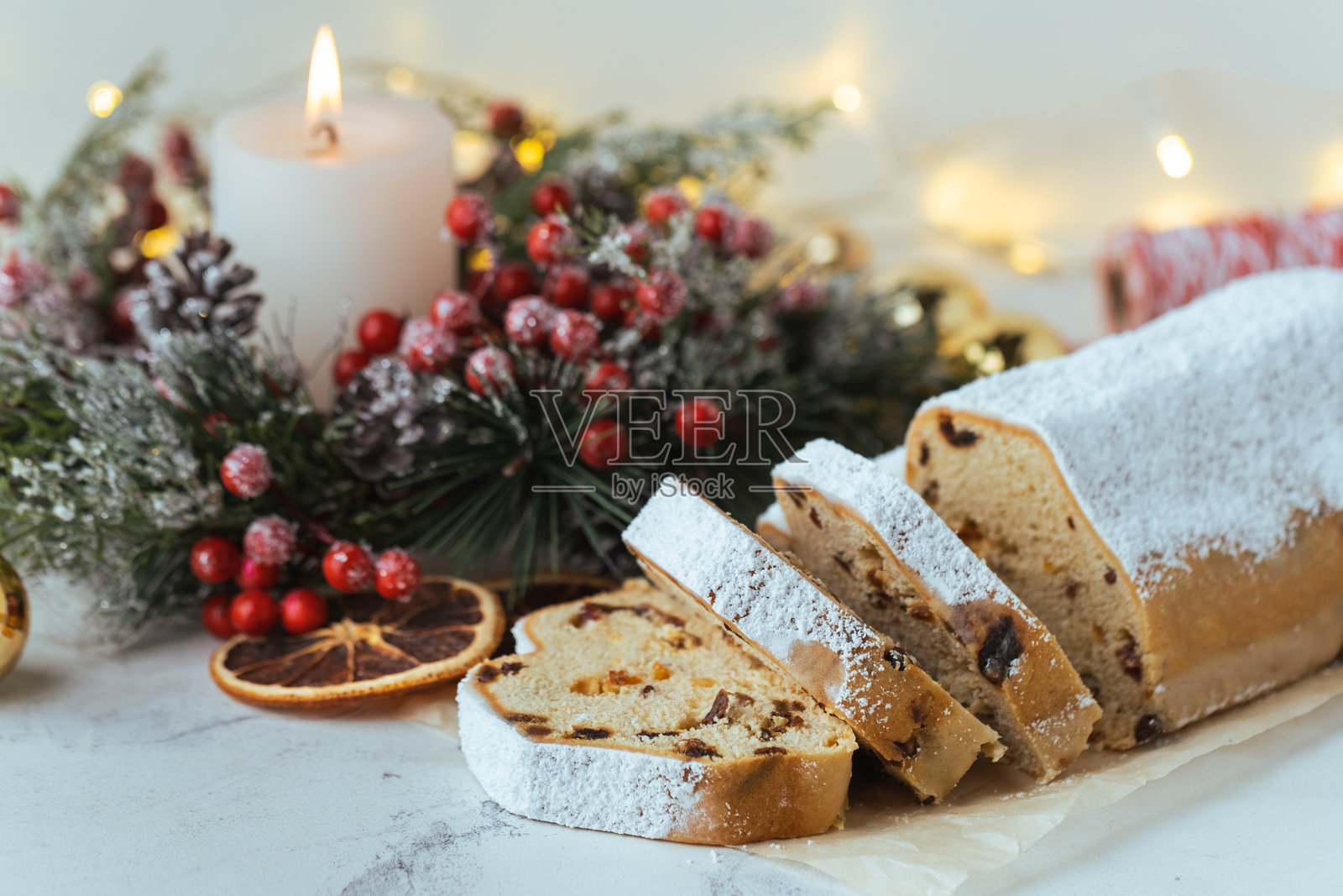 传统德国圣诞蛋糕斯托伦，内含葡萄干、水果和坚果，表面撒有糖粉，摆放在装饰有圣诞树枝、灯光和蜡烛的节日餐桌上。照片摄影图片