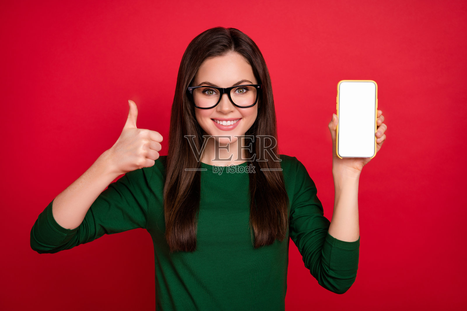 酷炫的棕发女老板展示电话，竖起大拇指，佩戴眼镜，穿着绿色衬衫，背景为红色。照片摄影图片